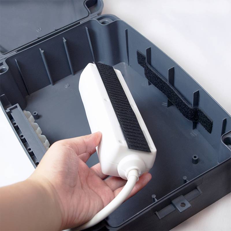 Una persona tiene un dispositivo elettronico rettangolare con un cavo, che viene posizionato in un involucro grigio aperto di plastica.