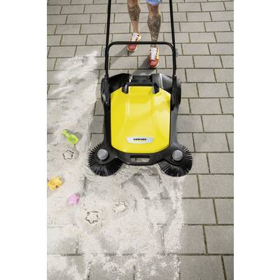 Una persona utilizza uno spazzaneve giallo su una superficie pavimentata, pulendo sabbia con stampi di giocattoli e impronte di piedi visibili.