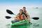 Due persone remano sorridenti in un kayak su acque blu e tranquille, entrambe indossano giubbotti di salvataggio.