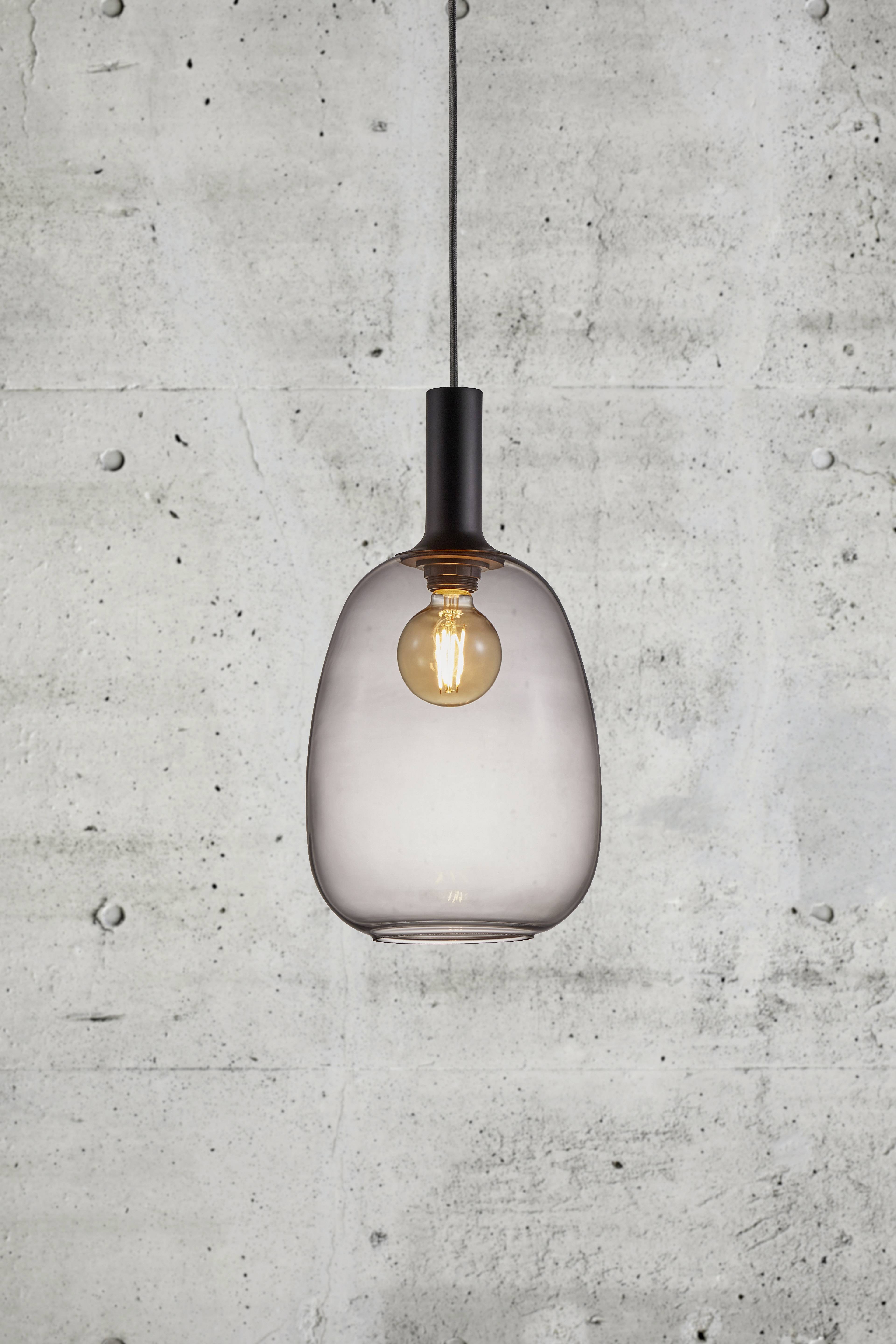 Una lampadina sospesa con paralume in vetro fumé davanti a un muro di cemento grigio.