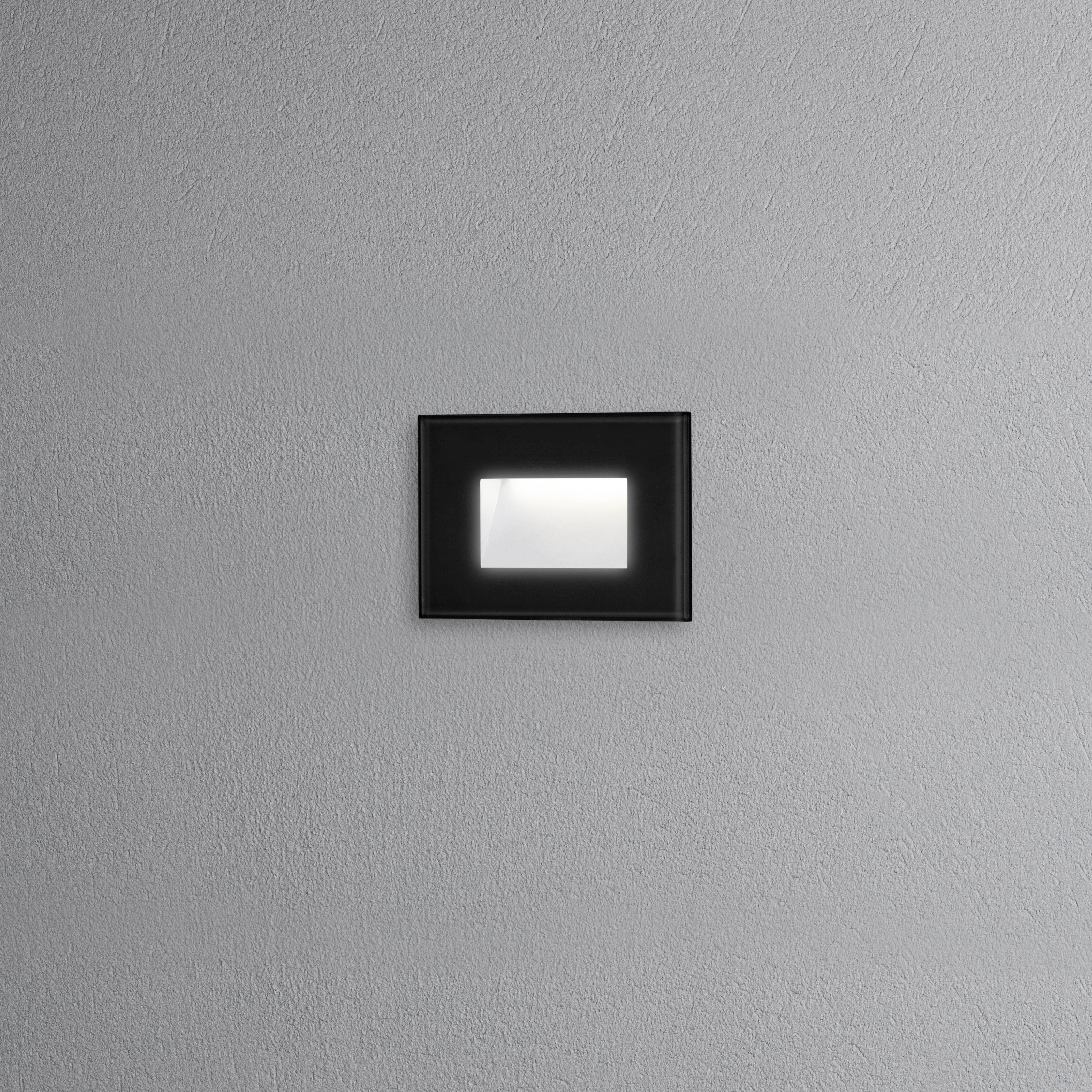Un'illuminazione murale nera e quadrata su una parete grigia con struttura, che illumina delicatamente l'ambiente circostante.