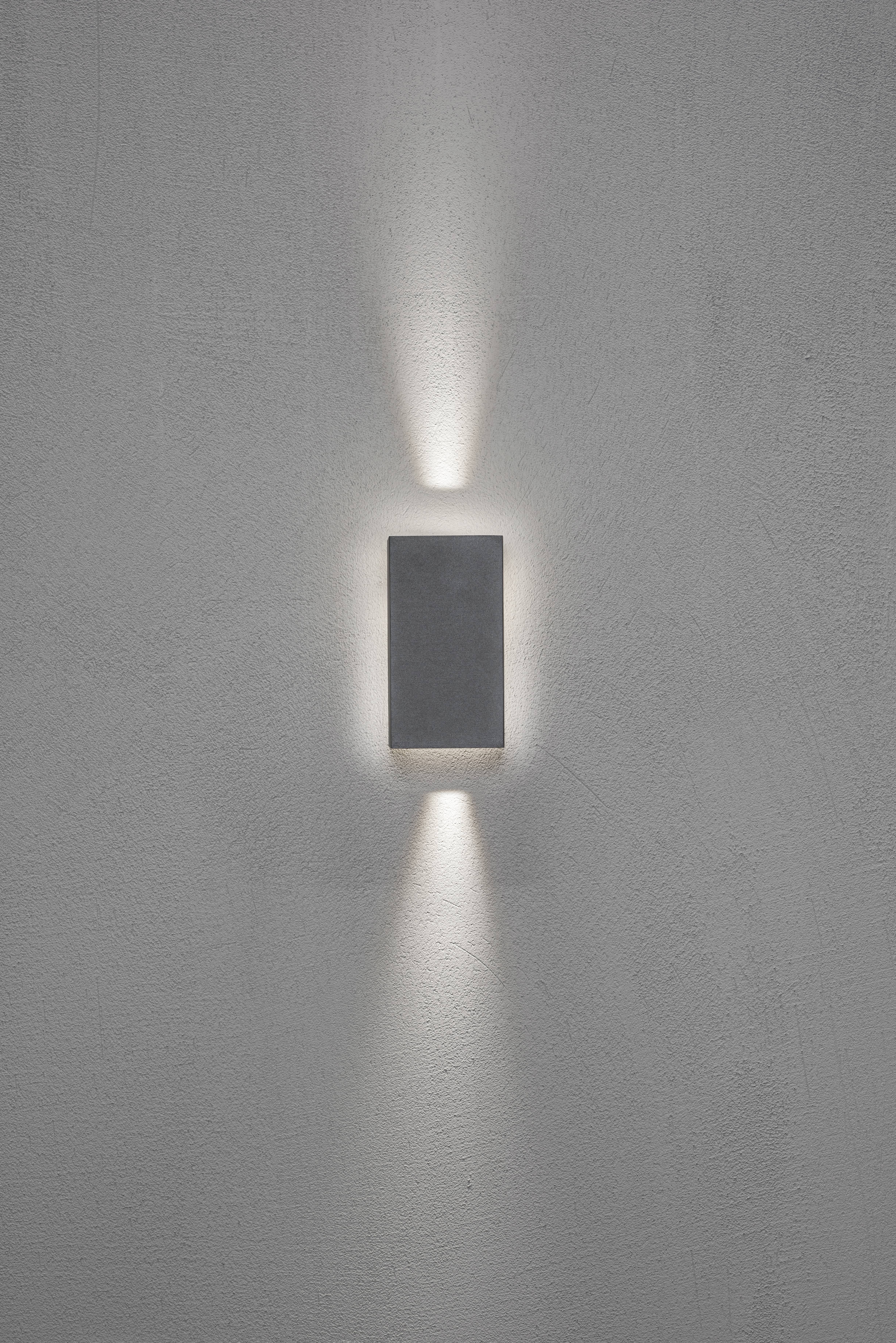 Una lampada da parete rettangolare illumina la parete verso l'alto e verso il basso, creando un delicato motivo di luce su uno sfondo grigio.
