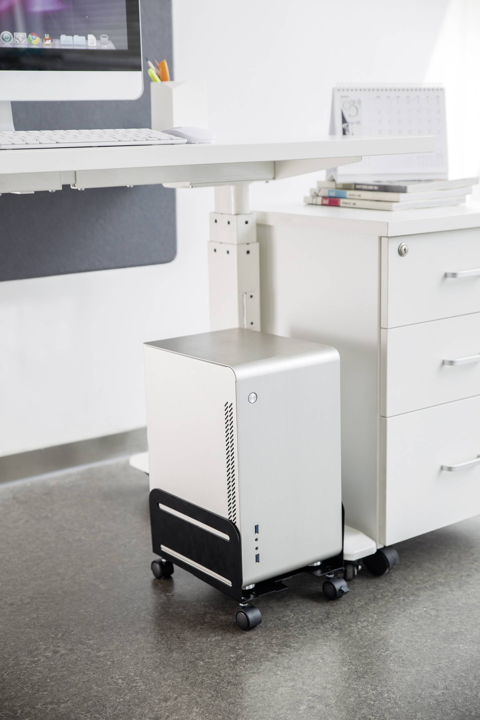 Un computer moderno bianco è posizionato su un carrello sotto una scrivania bianca, accanto a un mobile bianco con dei libri.