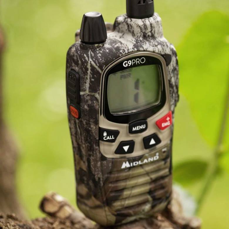 Un walkie-talkie mimetico, modello Midland G9PRO, è posizionato su un tronco d'albero nel verde. Mostra uno schermo vuoto.
