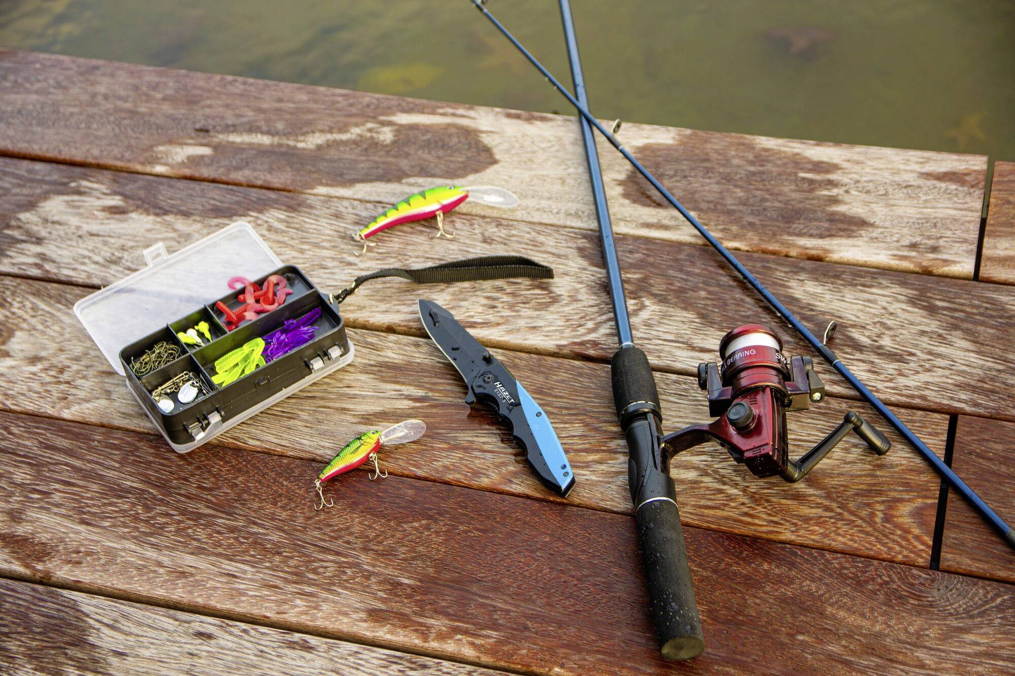 Attrezzatura da pesca su un pontile di legno: Due canne da pesca, una scatola per esche con esche colorate e un coltello pieghevole nero.