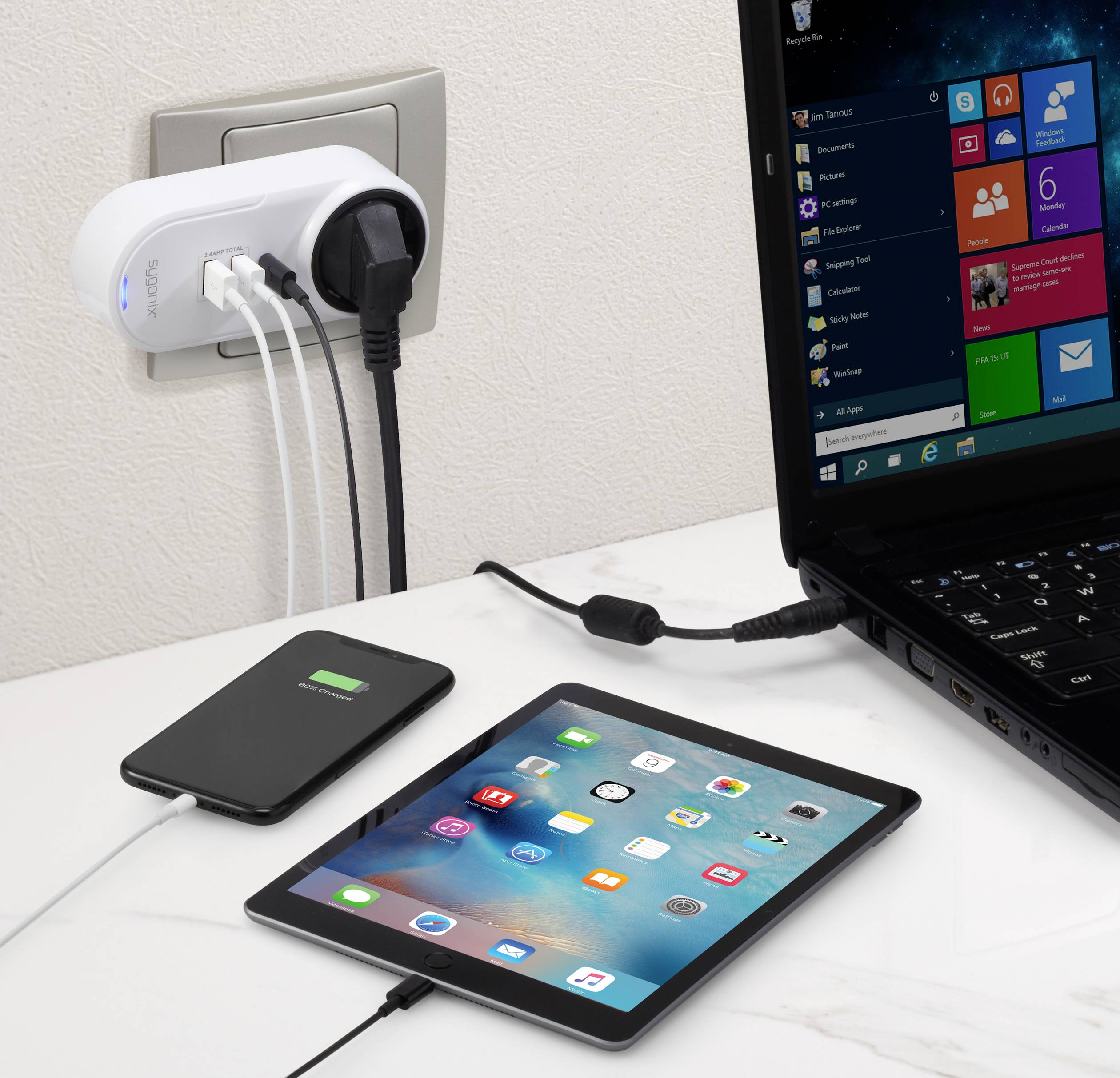 Un caricabatterie sta caricando un laptop, un tablet e uno smartphone su un tavolo. L'adattatore è collegato alla presa a muro.