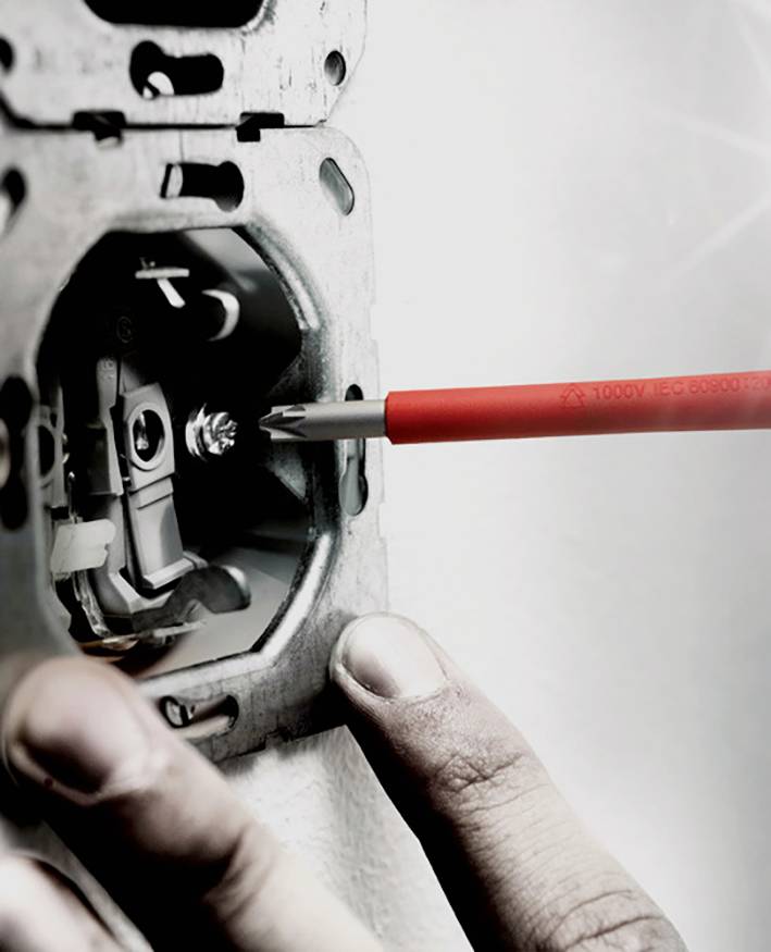 Una mano impugna un cacciavite mentre lavora su una presa elettrica, con focus sullo strumento e sulla presa, sullo sfondo sfocato.