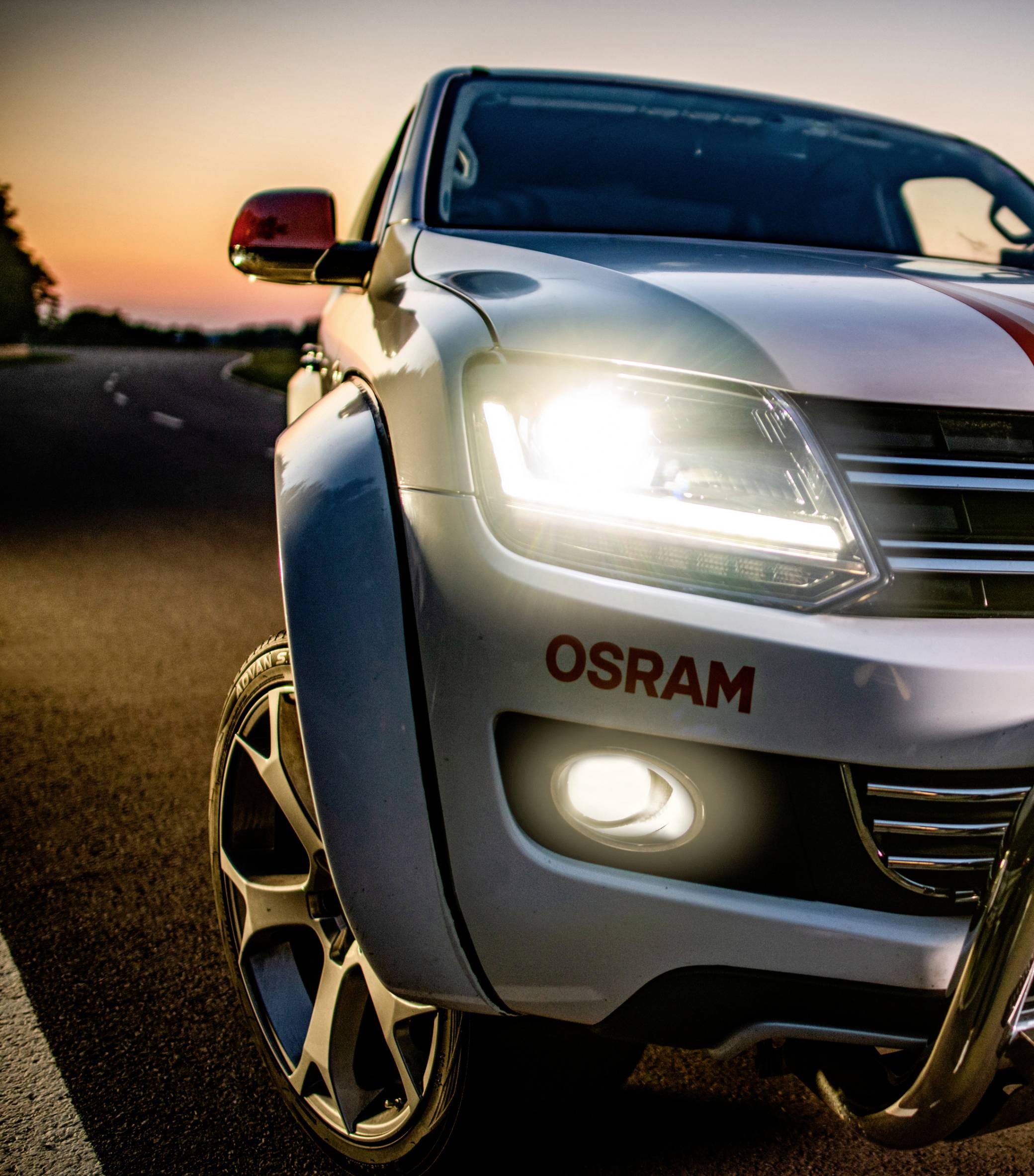 Un SUV bianco con i fari accesi è fermo su una strada al tramonto. Il nome del marchio 'OSRAM' è visibile sul paraurti.