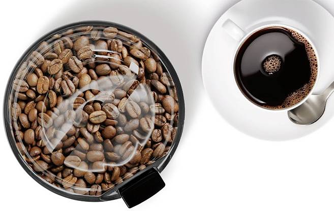 Chicchi di caffè in un contenitore rotondo per la conservazione, accanto a una tazza bianca con caffè appena preparato e un cucchiaio.