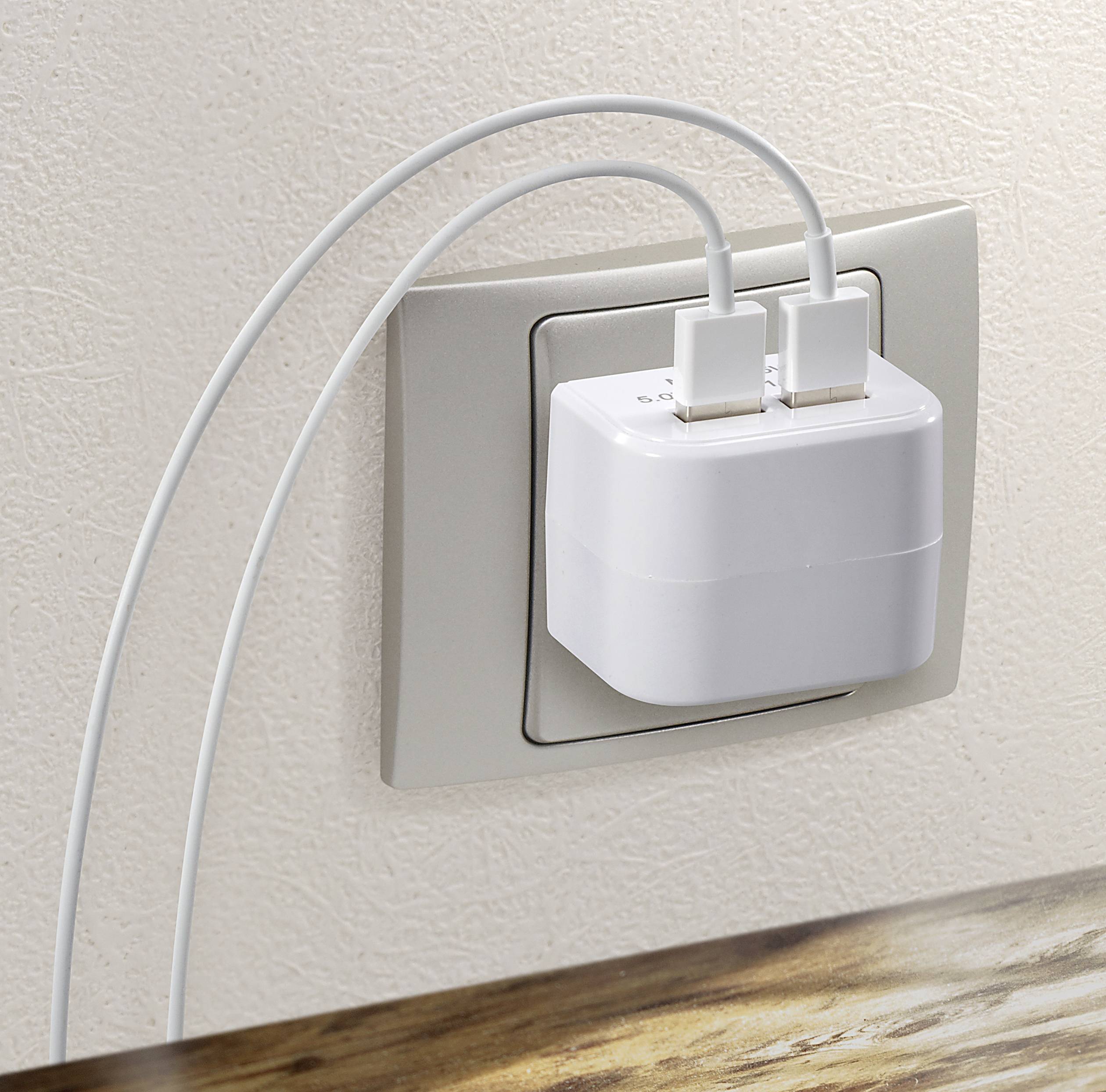 Un caricabatterie con due cavi USB collegati è inserito in una presa a muro, su un tavolo di legno in primo piano.