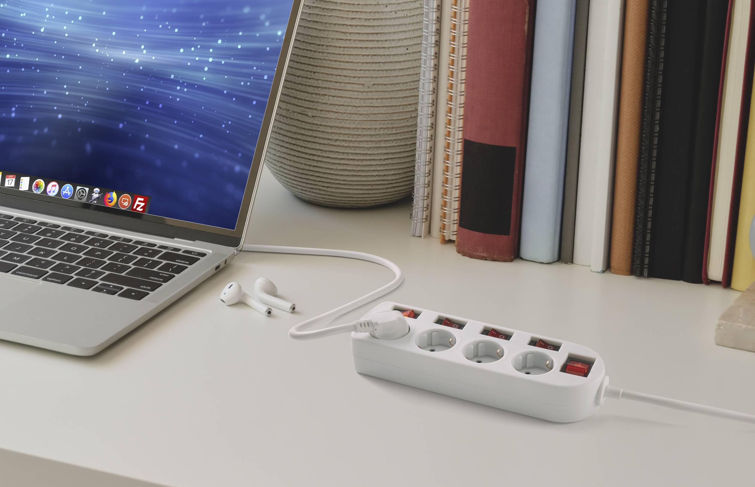 Un laptop su un tavolo è collegato a delle cuffie. Accanto, è posizionata una multipresa con quattro prese davanti ad alcuni libri.