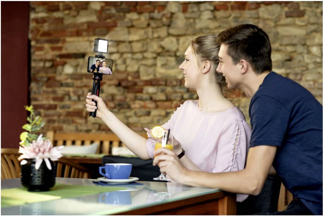 Una coppia in un caffè scatta un selfie con uno smartphone su un treppiede. Sorridono, e ci sono delle bevande sul tavolo.
