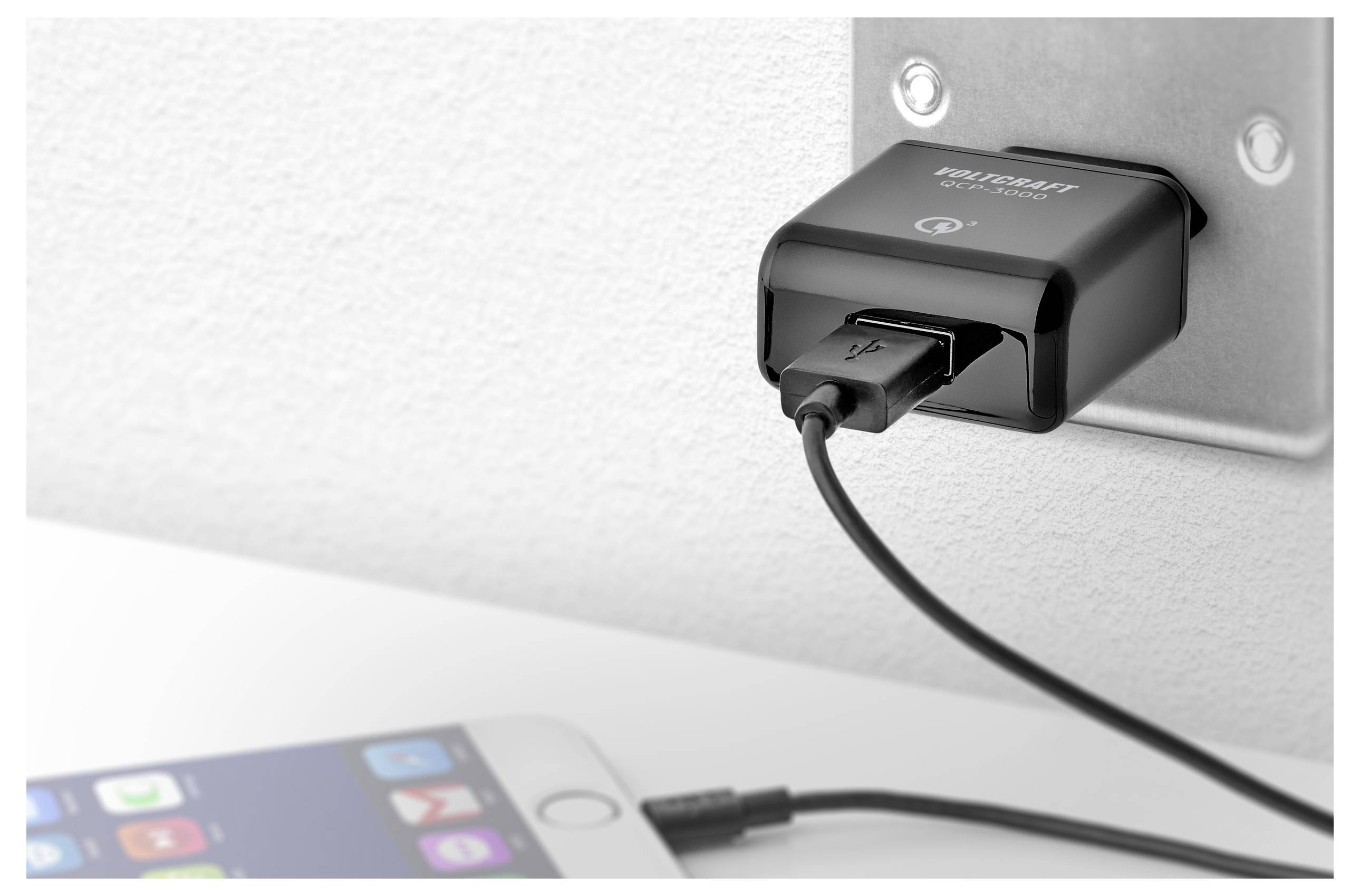 Un caricabatterie USB nero inserito in una presa a muro sta caricando uno smartphone su un tavolo bianco in primo piano.