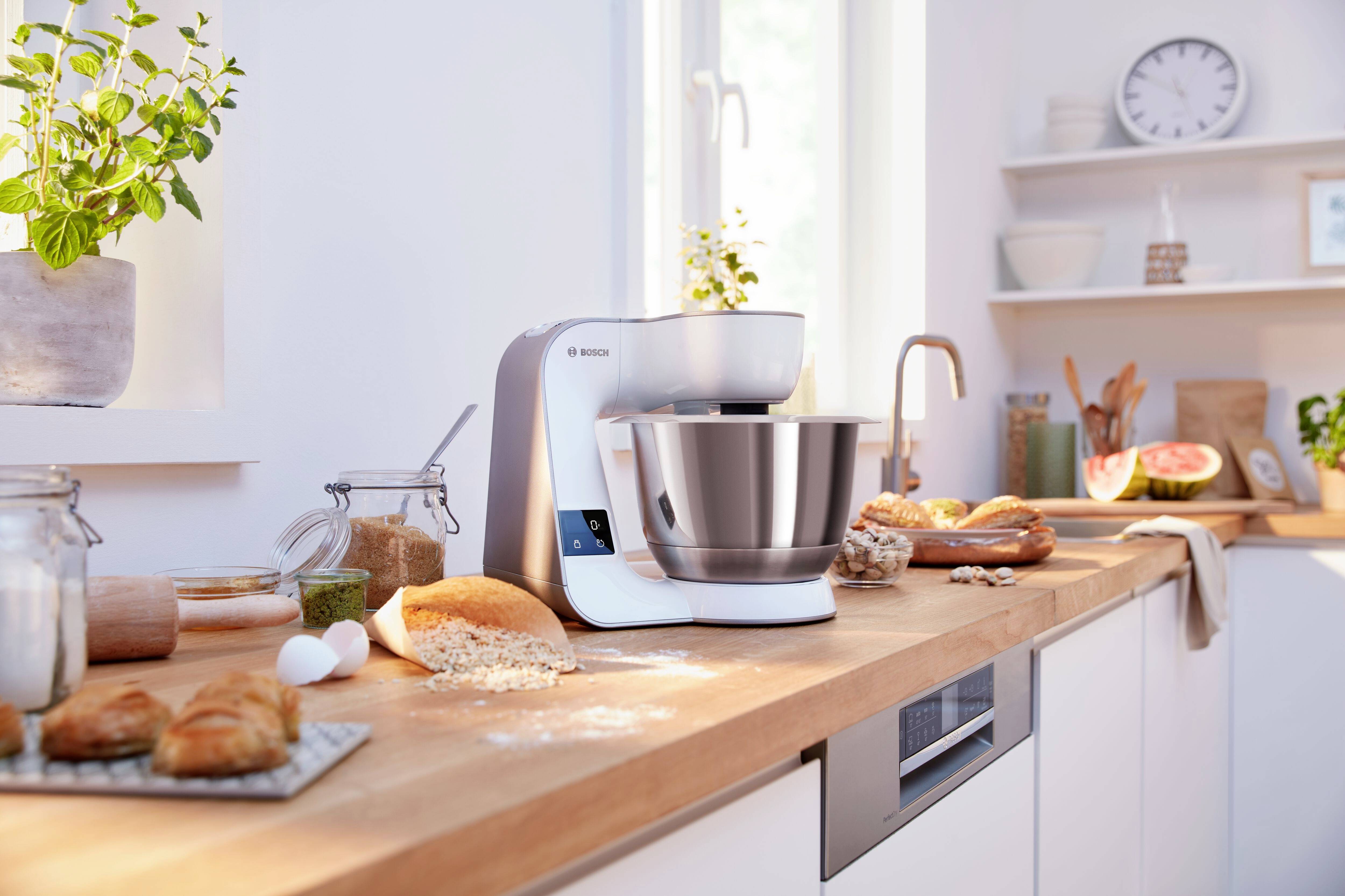 Moderna macchina da cucina su una superficie di lavoro bianca, circondata da ingredienti freschi come farina, uova e pane. Cucina luminosa e arieggiata.
