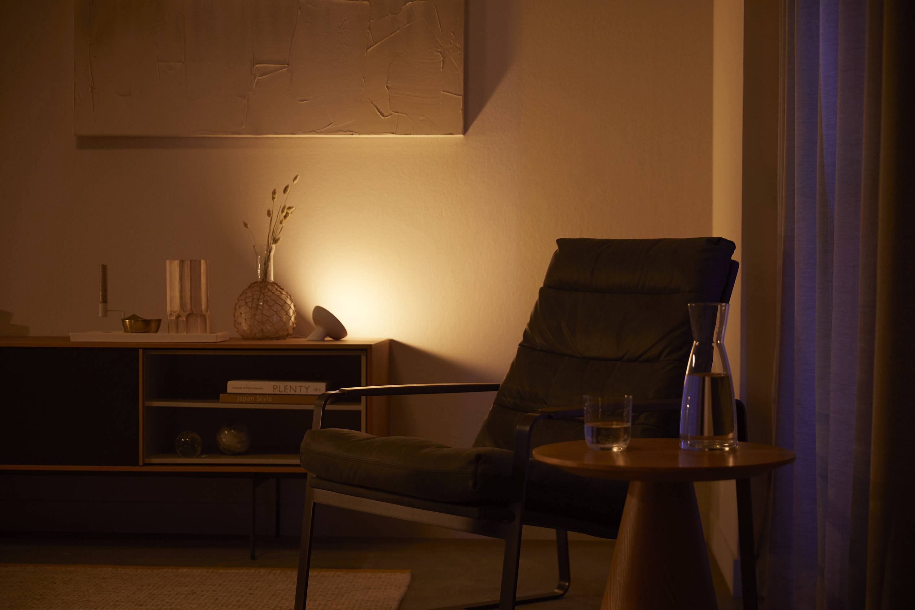 Un ambiente accogliente con una poltrona, un tavolino con un bicchiere e una caraffa. L'illuminazione di sfondo crea un'atmosfera calda.