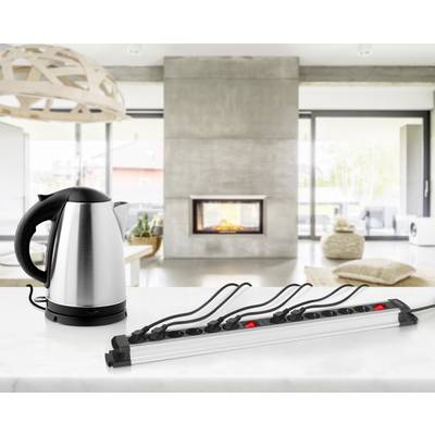Un bollitore è posizionato su un piano di lavoro della cucina; accanto c'è una ciabatta elettrica lunga. Sullo sfondo è visibile un ambiente domestico moderno con un camino.