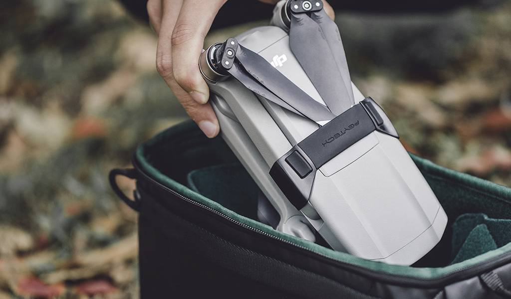 Una mano sta inserendo un drone in una borsa nera. L'ambiente è ricoperto di foglie.