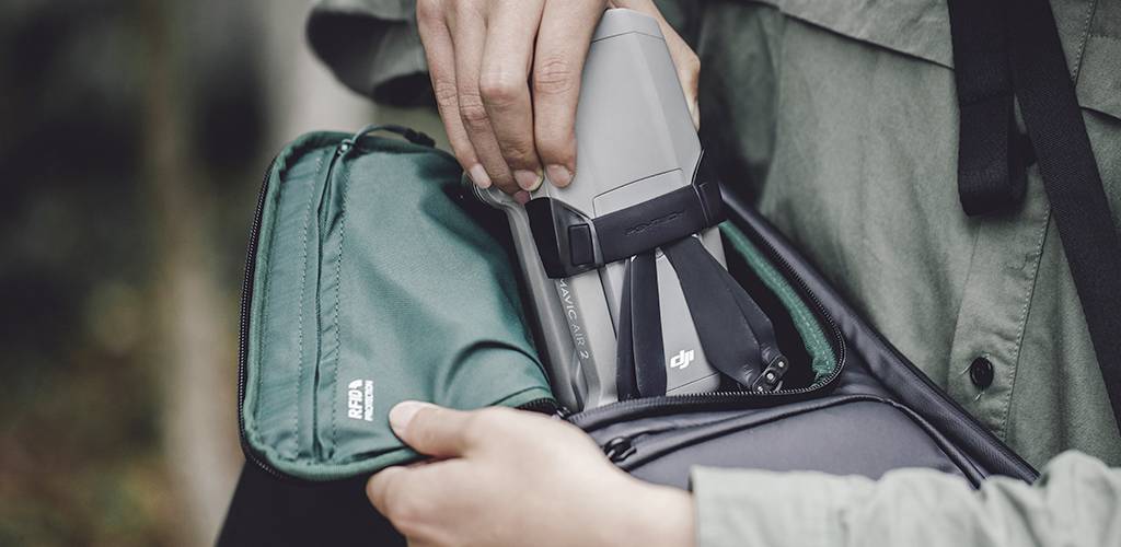 Una persona sta inserendo un drone grigio in una borsa verde. La scena suggerisce un'attività all'aperto. Le mani tengono la borsa aperta.