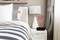Una camera da letto con un comodino, su cui si trovano una lampada e una sveglia digitale che mostra l'ora 12:08 e la temperatura di 87,5°F.