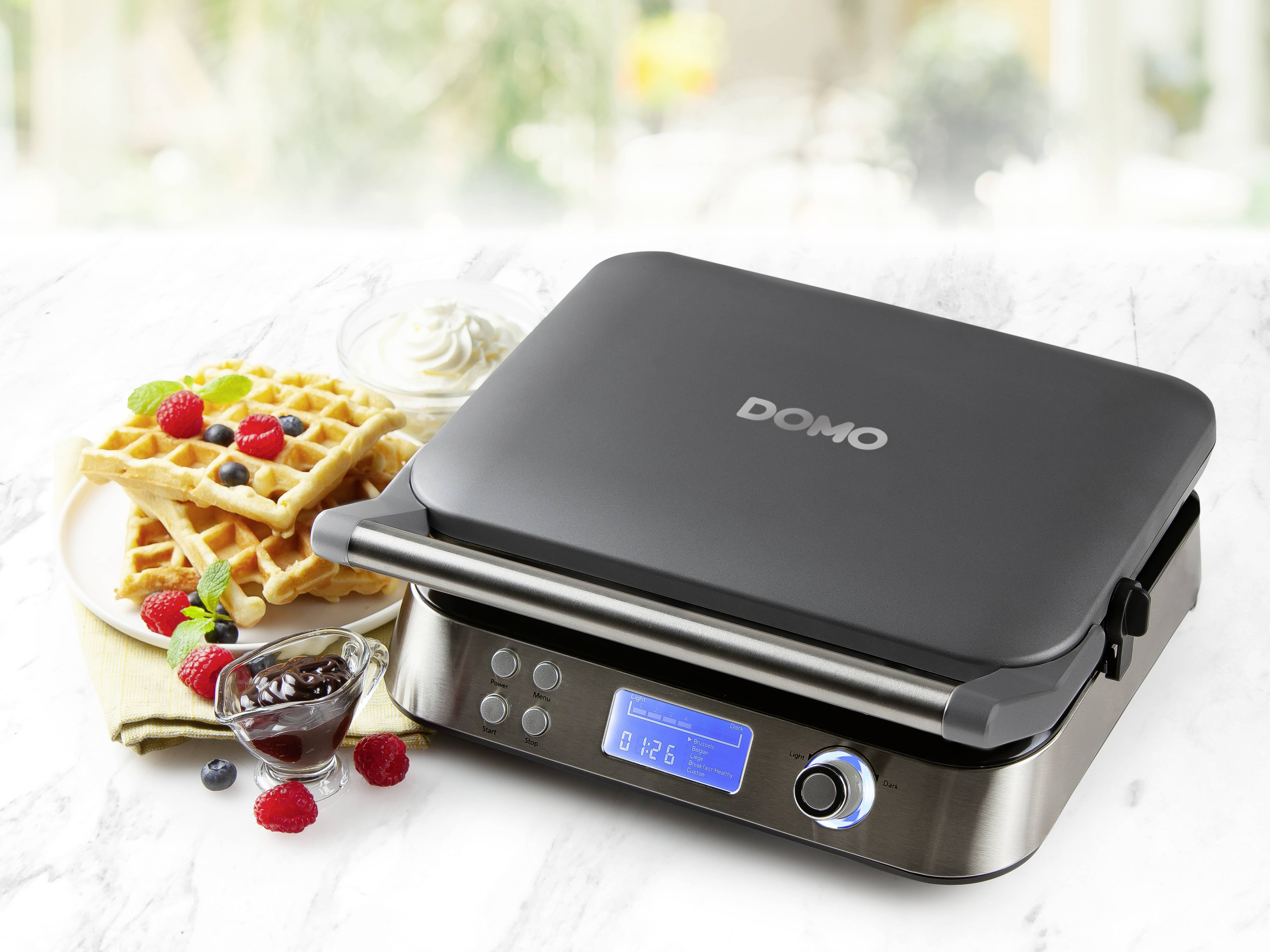 Macchina elettrica per waffle con display digitale sul piano della cucina. Accanto ci sono waffle appena sfornati con frutti di bosco e panna.