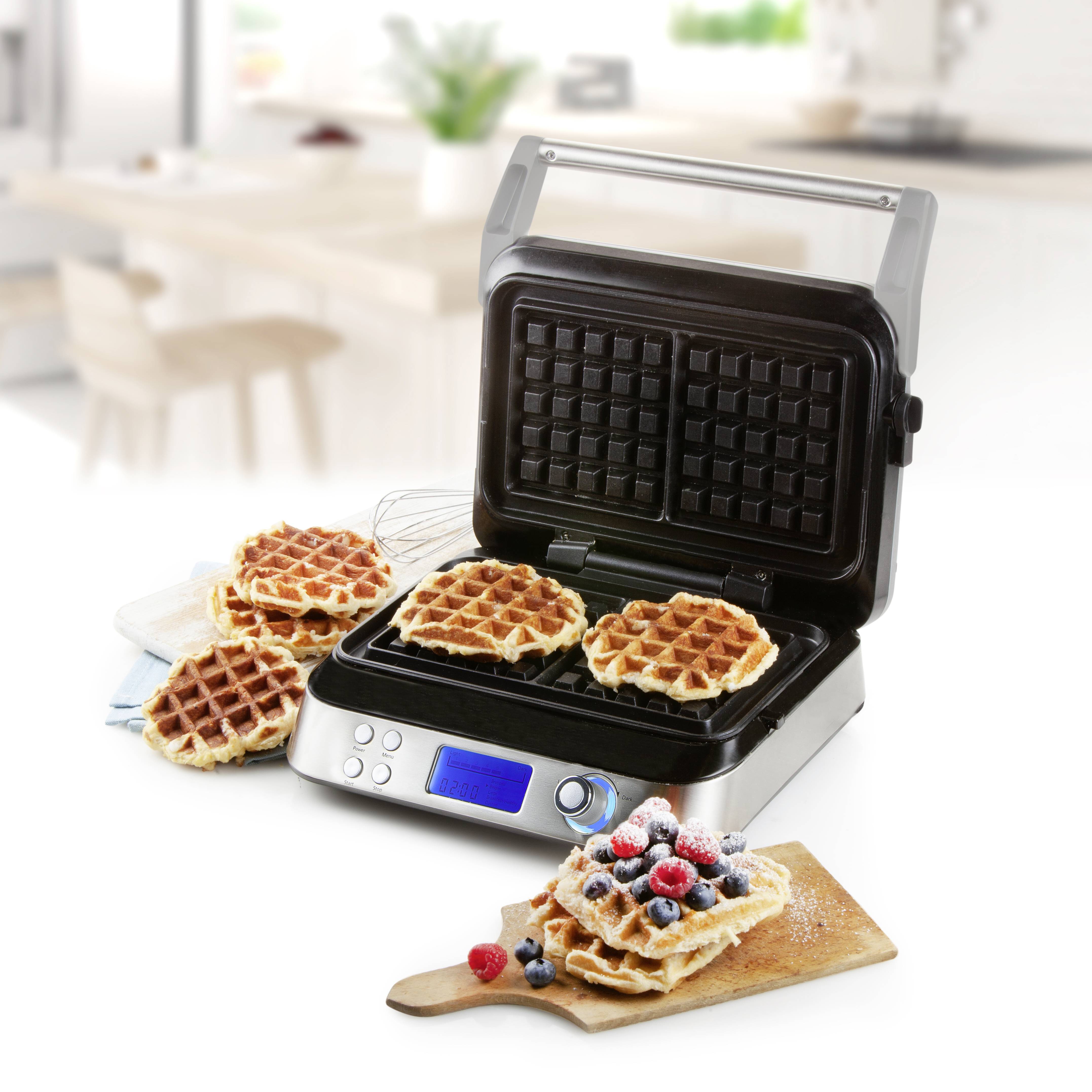 Macchina per waffle con coperchio aperto, con waffle appena cotte all'interno. Accanto un tagliere di legno con una waffle e bacche. Sullo sfondo: cucina.