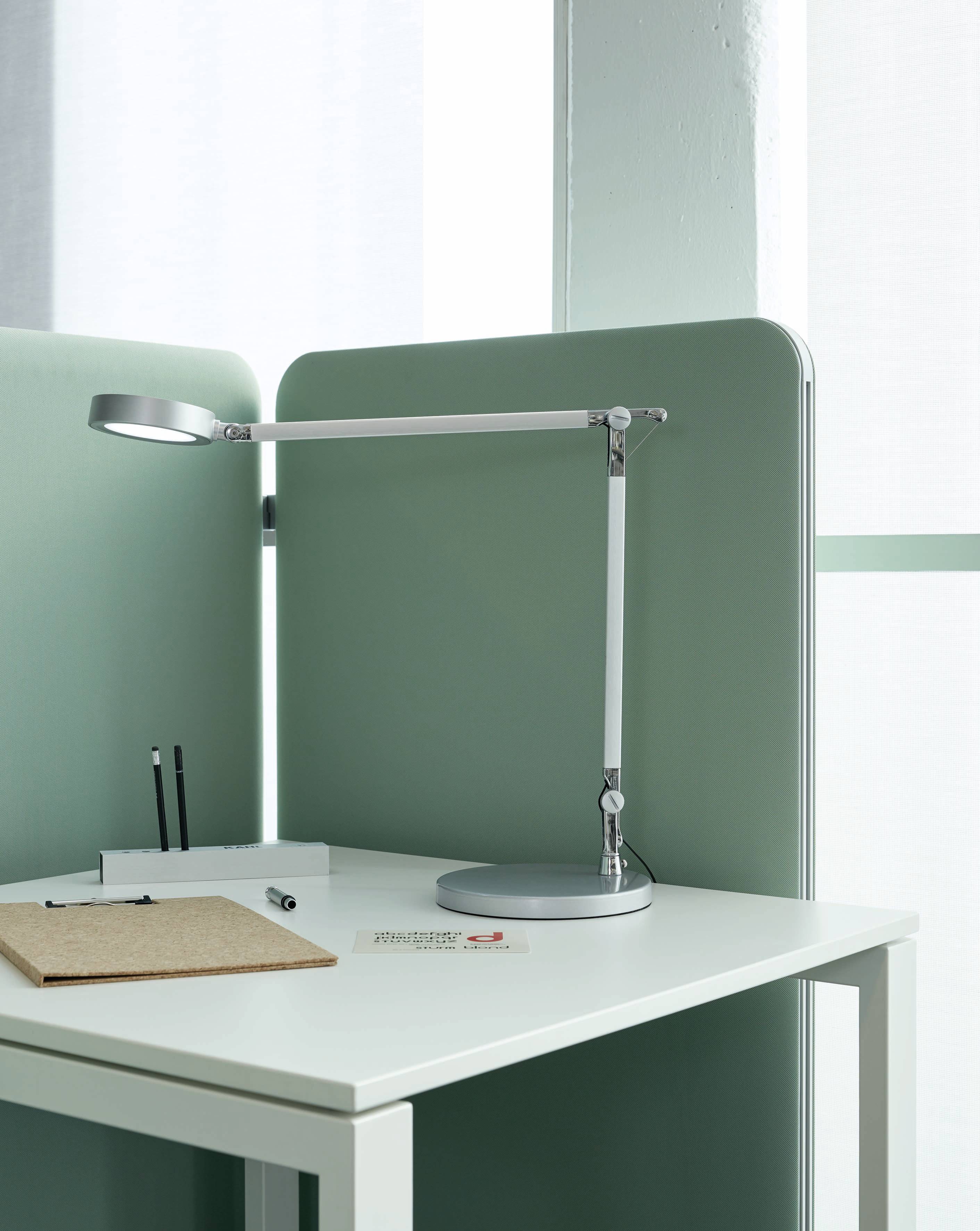 Una scrivania moderna con pannello divisorio verde, lampada da scrivania argentata, penne, blocco per appunti e biglietti da visita sul tavolo bianco.