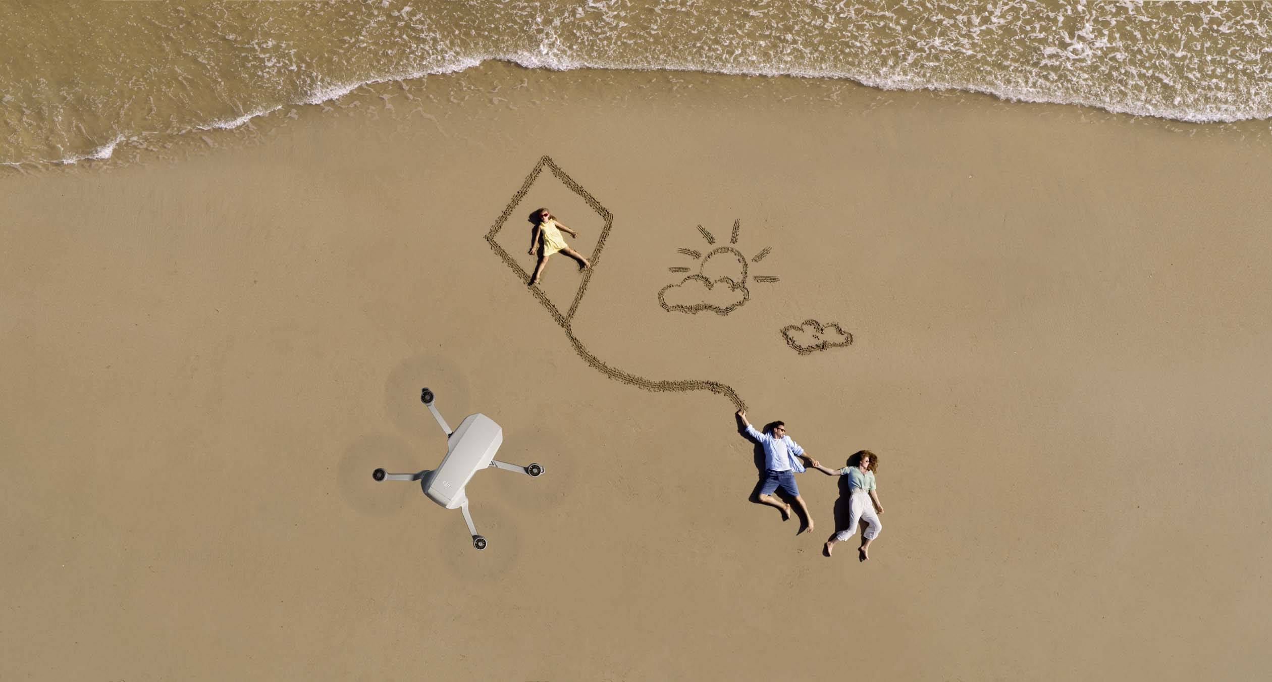 Una coppia è sdraiata sulla spiaggia, tiene un drone disegnato come un aquilone, con un bambino nell'aquilone. Accanto a loro è disegnato un sole e due nuvole nella sabbia.