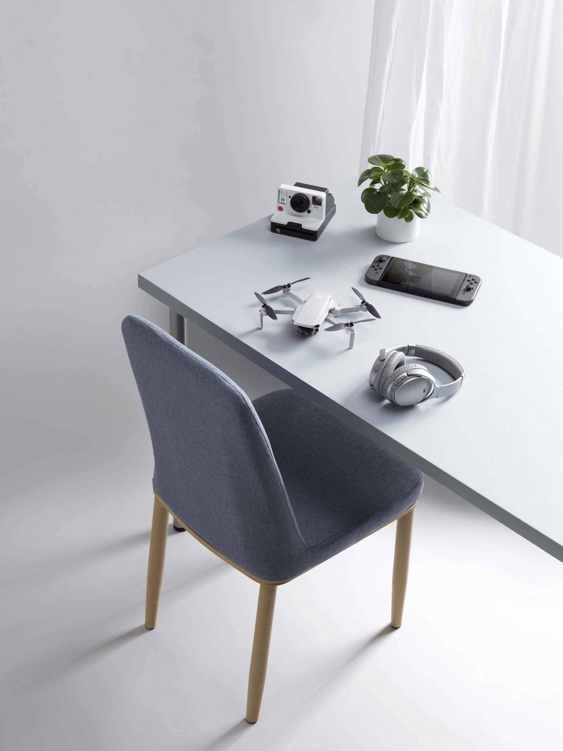 Su un tavolo ci sono un drone, una fotocamera, un dispositivo portatile per giocare e un paio di cuffie. Accanto si trovano una piccola pianta e una sedia.