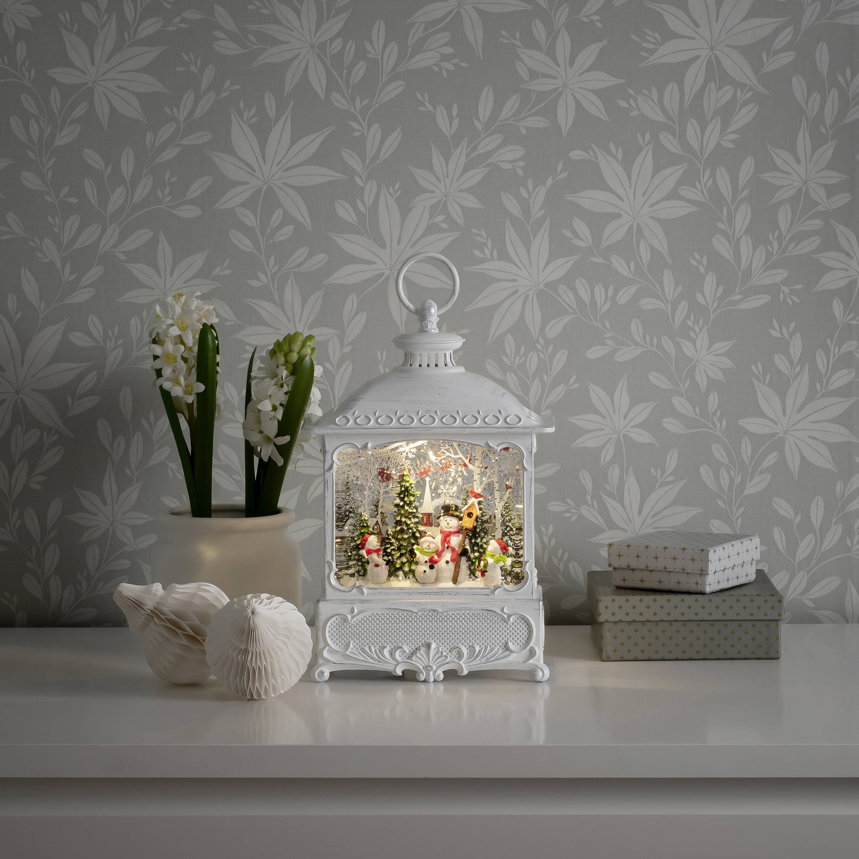 Una lanterna decorativa con una scena festiva è posizionata su un tavolo bianco accanto a due scatole regalo e un vaso di fiori.