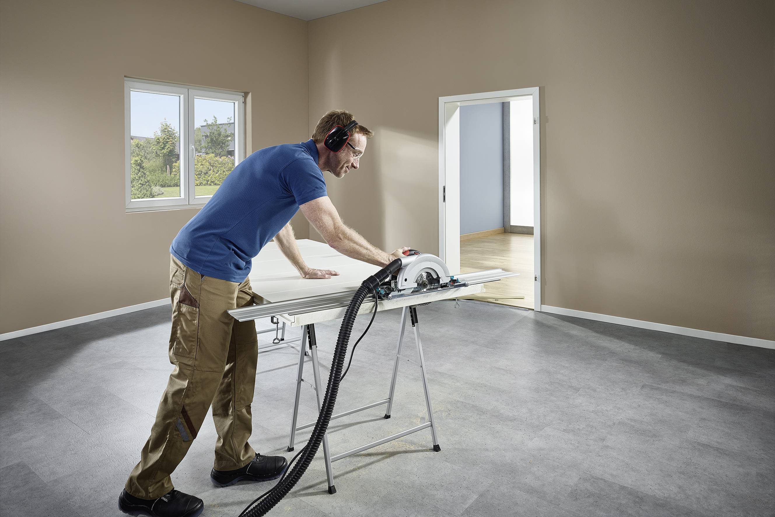 Un uomo sta usando una sega circolare per tagliare legno su un banco da lavoro in una stanza vuota. Indossa protezioni per l'udito e abiti da lavoro.