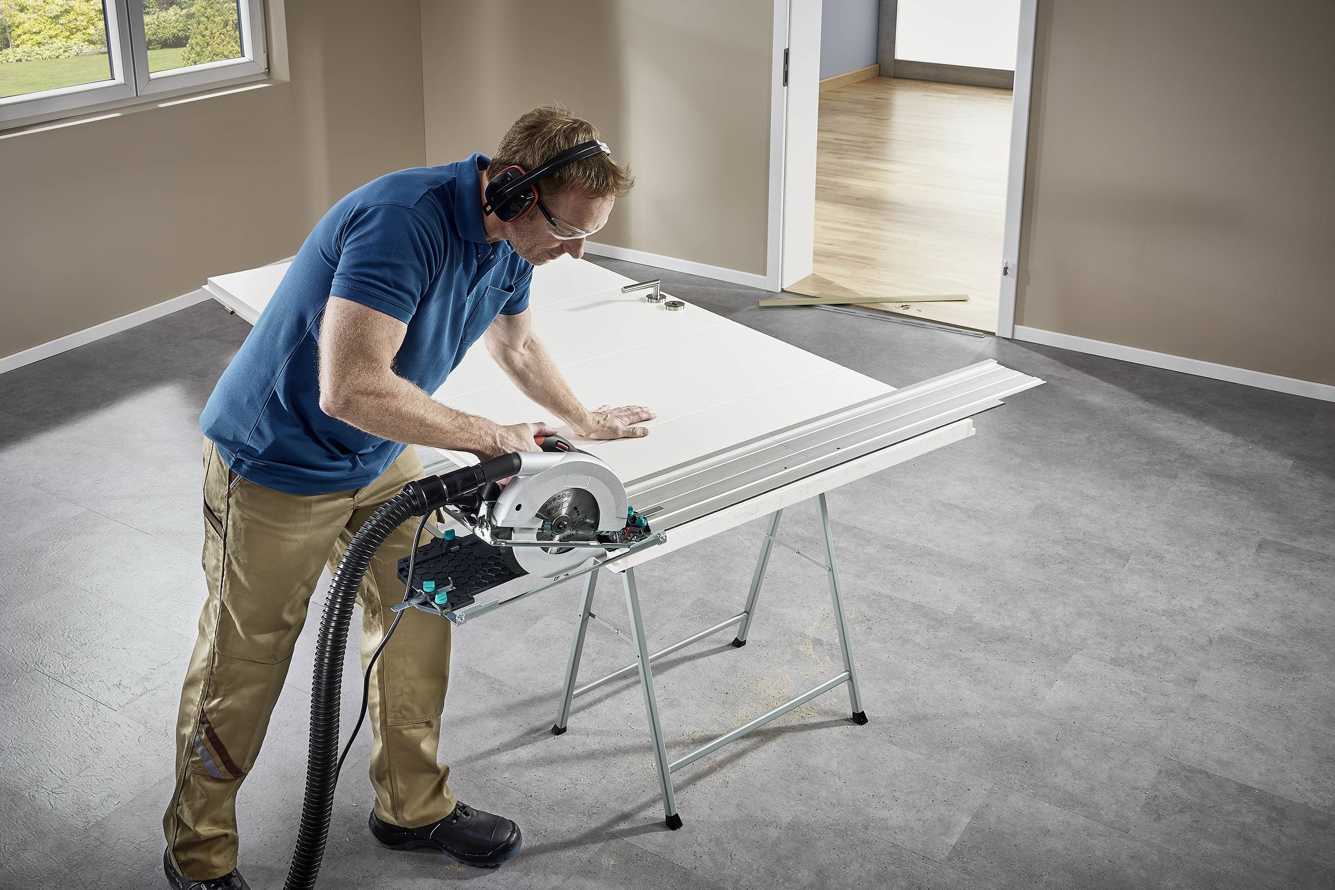 Un uomo con occhiali protettivi e cuffie antirumore taglia una grande tavola di legno con una sega circolare in un ambiente ben illuminato.