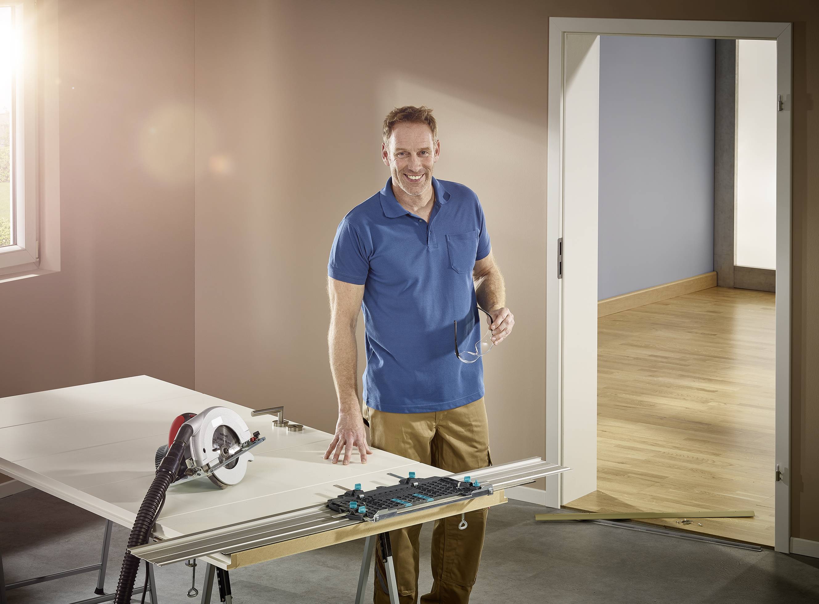 Un uomo sorridente con una polo blu sta lavorando in una stanza con una sega circolare su un tavolo accanto a lui. Si trova in un ambiente ben illuminato con un pavimento in legno.