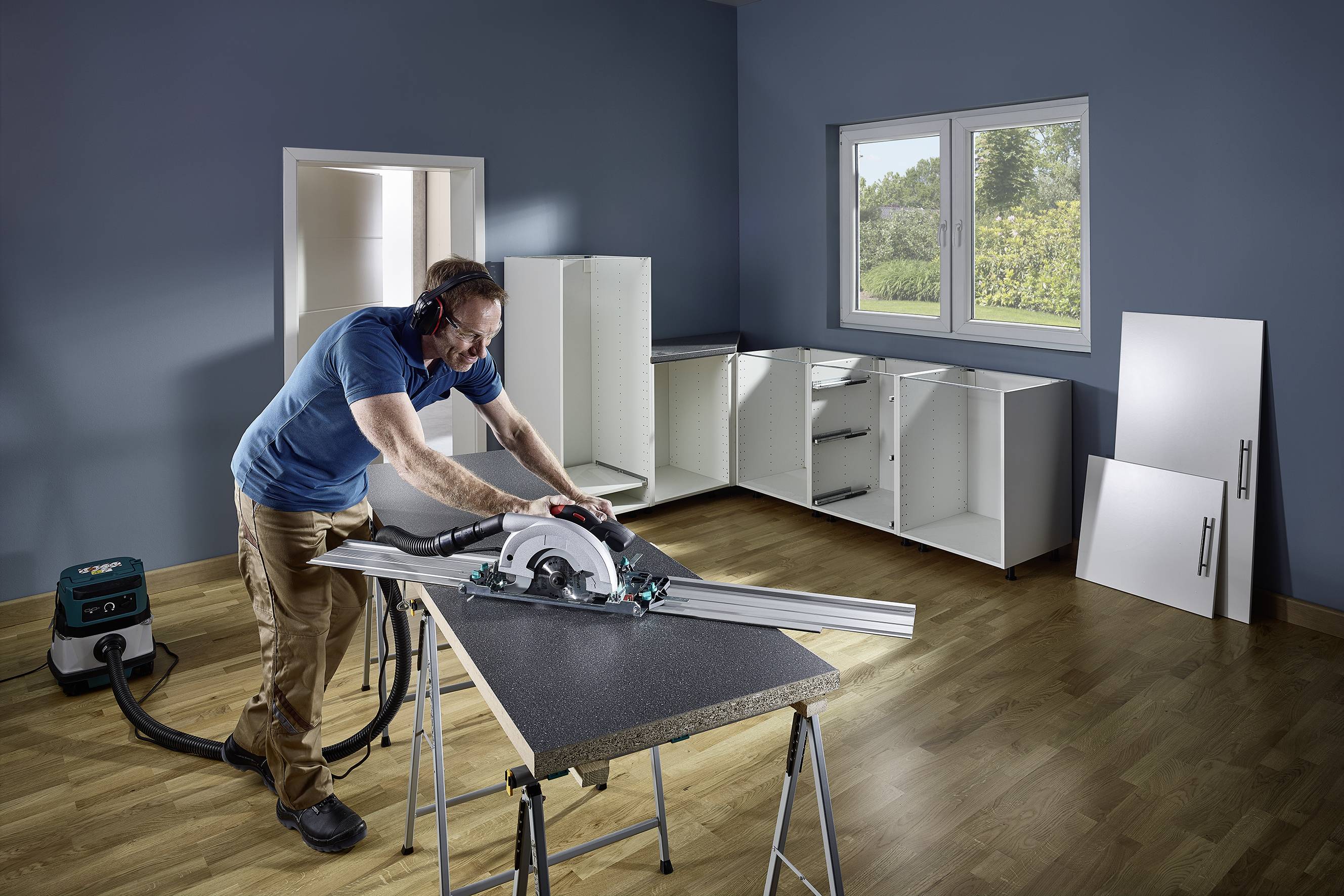 Un uomo sta lavorando con una sega circolare in una stanza con pareti dipinte di blu, mentre assembla gli armadi da cucina.