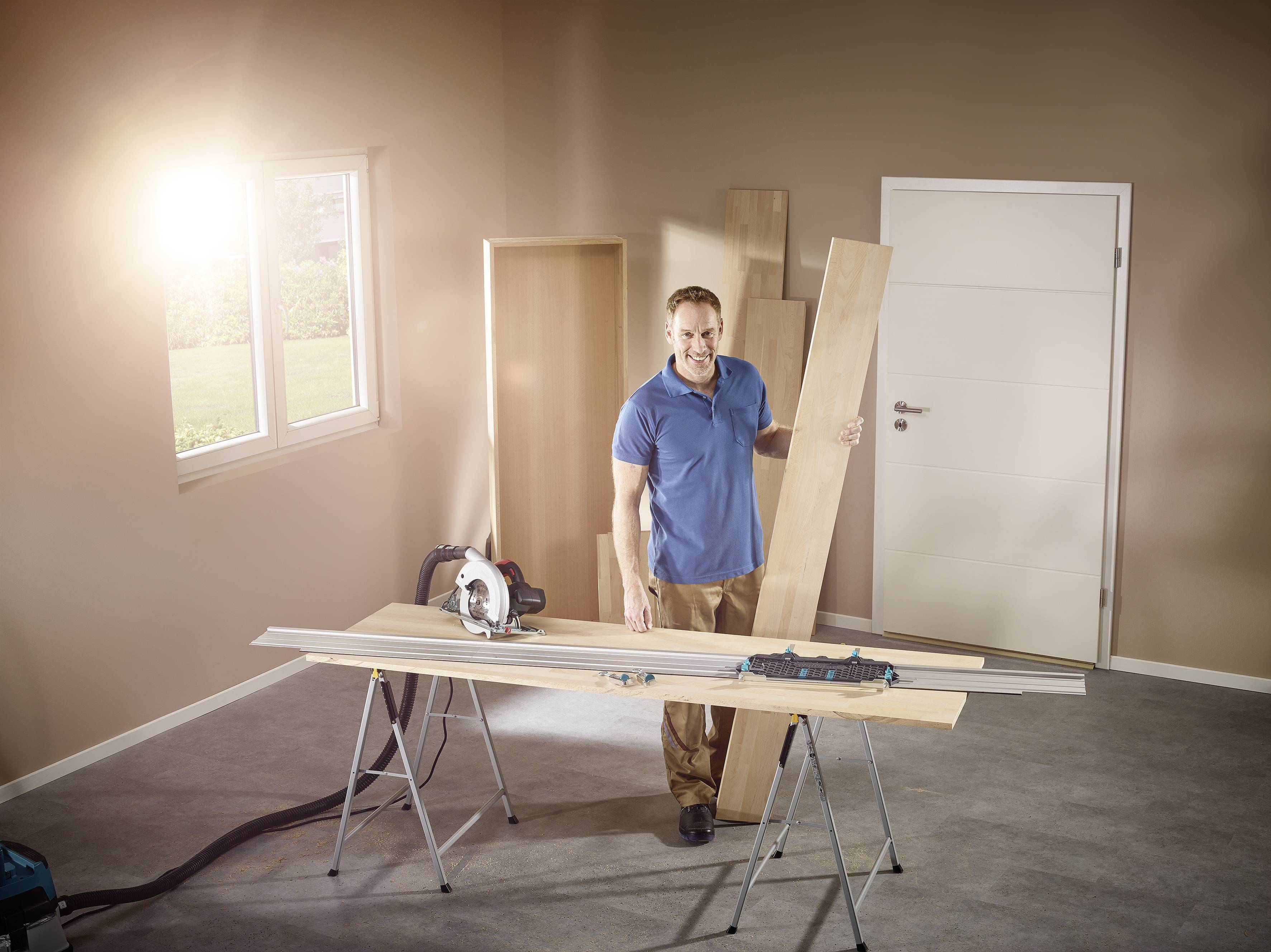 Un uomo in un locale con materiale edile tiene in mano una tavola di legno. Sullo sfondo ci sono parti di legno non assemblate e una sega circolare su un tavolo.
