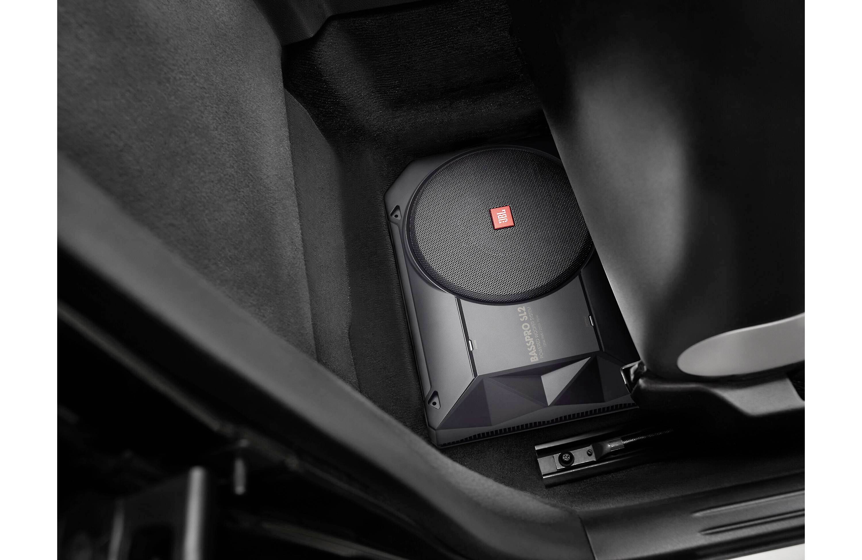 Un subwoofer è installato nel vano piedi di un veicolo, circondato da un tappeto scuro. Il design compatto massimizza lo spazio disponibile.