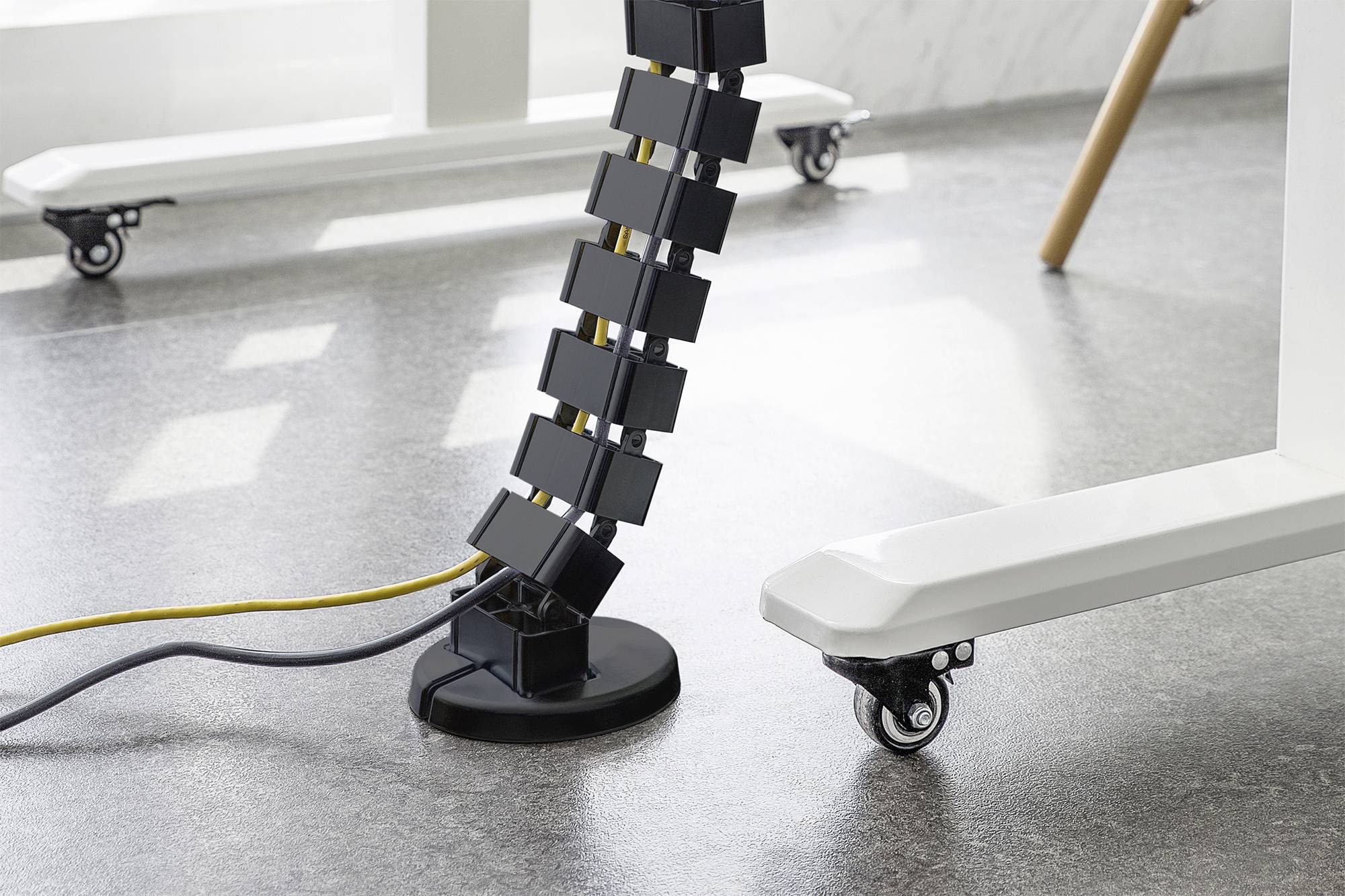 Una catena portacavi flessibile nera scende da un tavolo fino al pavimento. I cavi sono ordinatamente sistemati al suo interno. Gambe del tavolo con rotelle sono visibili.