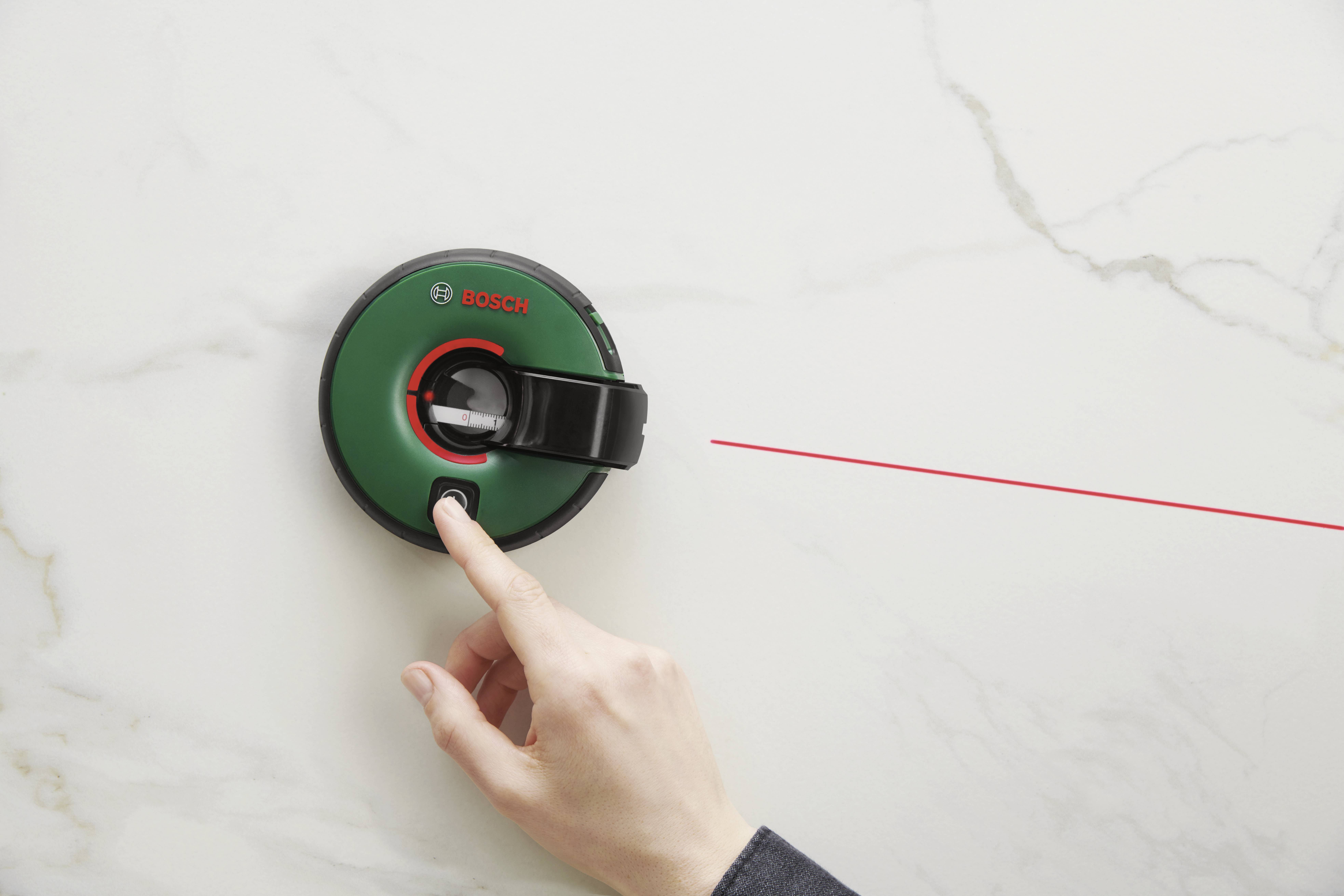 Una mano preme un misuratore di distanza laser verde che proietta un raggio laser rosso su una superficie bianca.