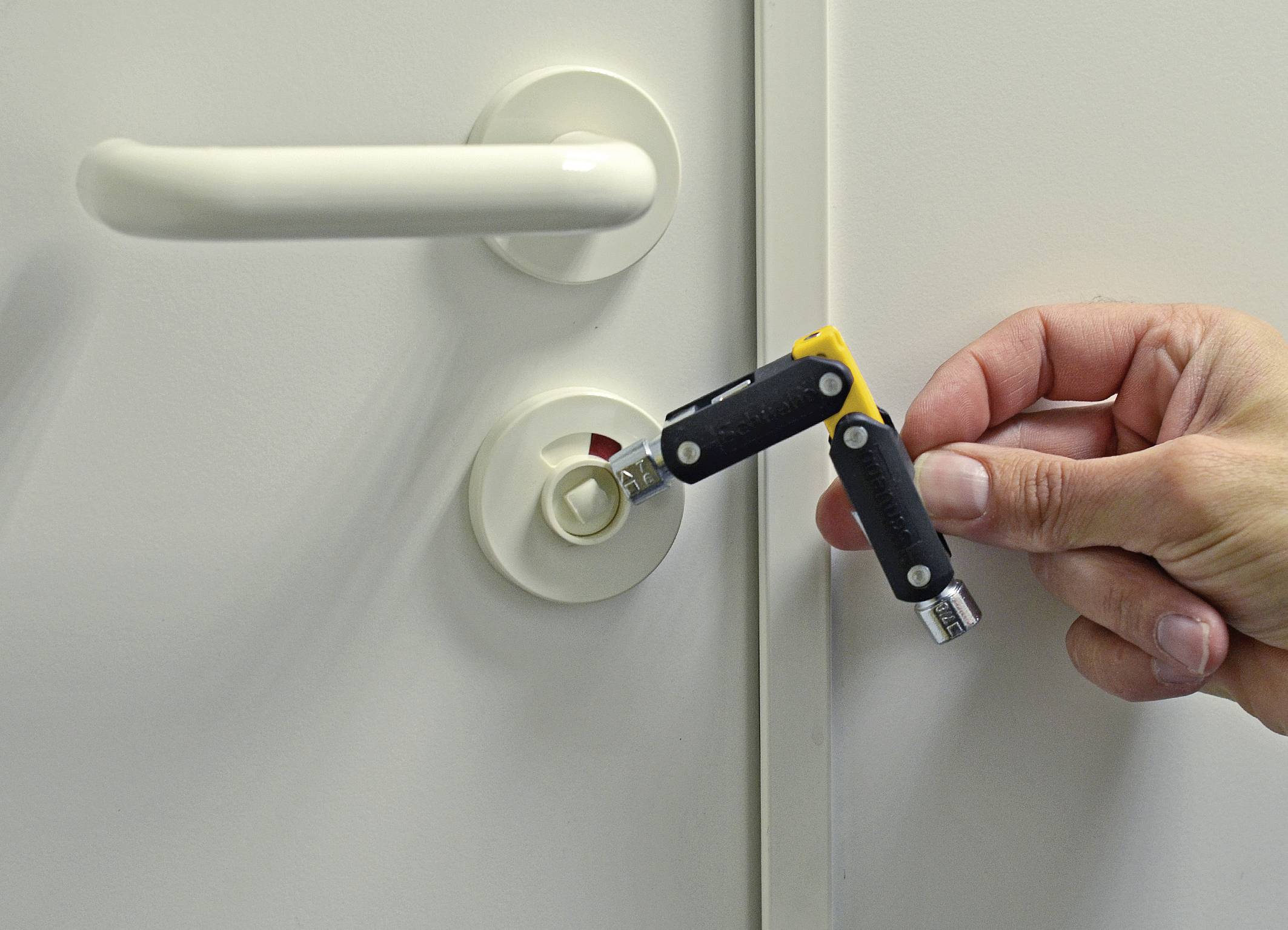 Una mano che tiene un attrezzo per forzare serrature vicino a una porta bianca con maniglia e serratura, rappresentazione di un rischio di sicurezza tramite strumenti per effrazione.