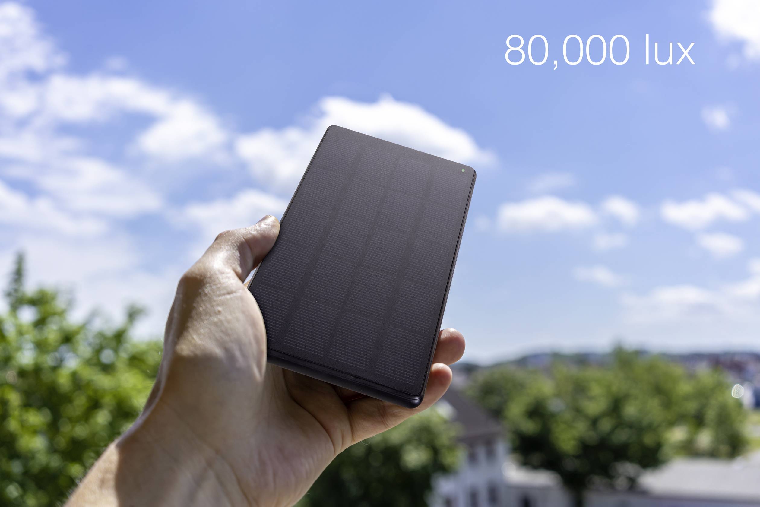 Mano che tiene una cella solare rettangolare scura all'aperto soleggiato; il testo mostra "80.000 lux", indicando un'elevata luminosità.