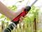 Una mano tiene un ugello per tubo da giardino davanti a piante verdi e una recinzione in legno. L'ugello è parzialmente rosso con un manico nero.
