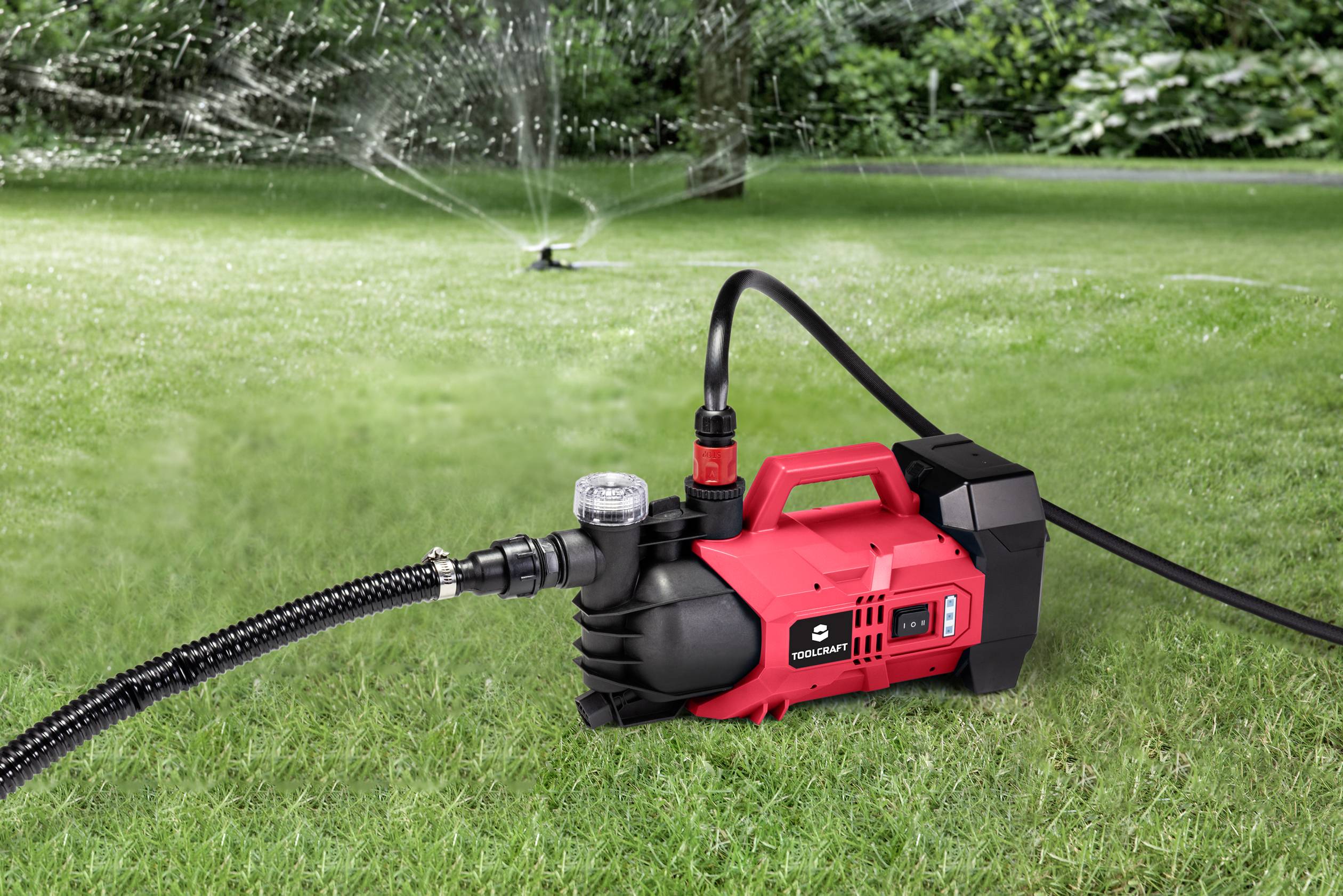 Pompa da giardino rossa su prato verde, spruzza acqua attraverso un tubo verso un irrigatore distante, con piante sullo sfondo.