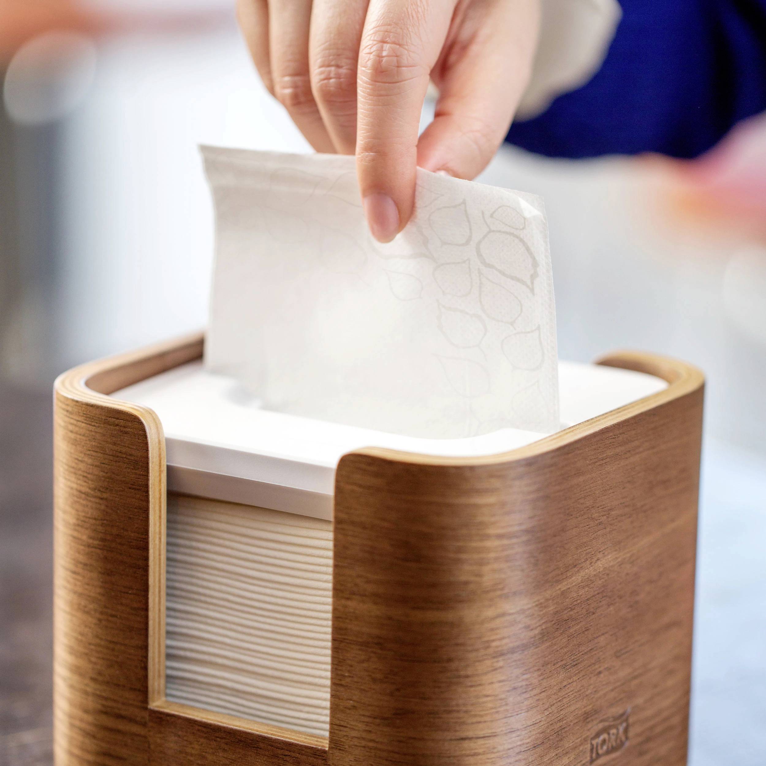 Una mano estrae un fazzoletto di carta da un dispenser di legno su un tavolo.