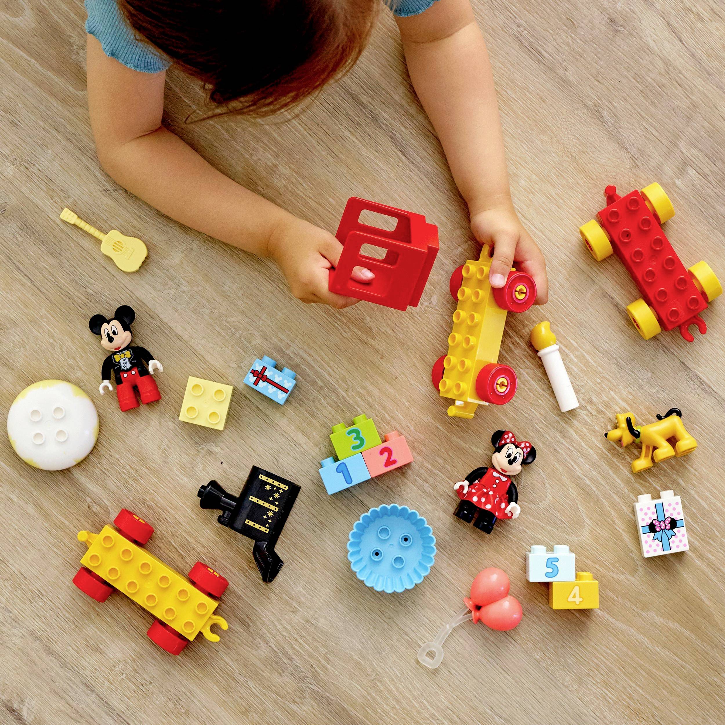 Un bambino gioca su un pavimento in legno con giocattoli colorati: un'auto Lego, figurine di Topolino e Minnie Mouse, regali, un palloncino e altro ancora.