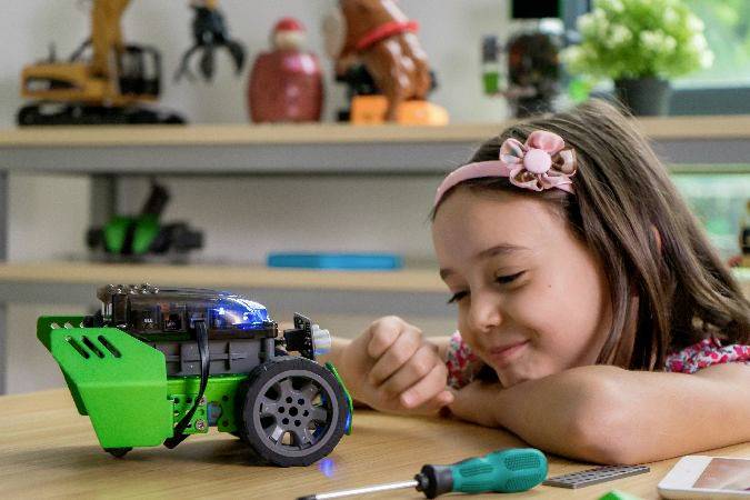 Una bambina osserva con gioia un piccolo robot verde su un tavolo. Attrezzi e pezzi sono sparsi accanto.