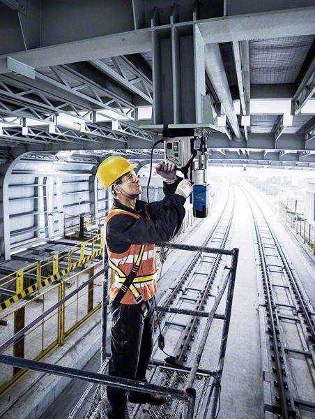 Un operaio edile con casco giallo e giubbotto di sicurezza lavora su un dispositivo tecnico sopra dei binari in un tunnel.