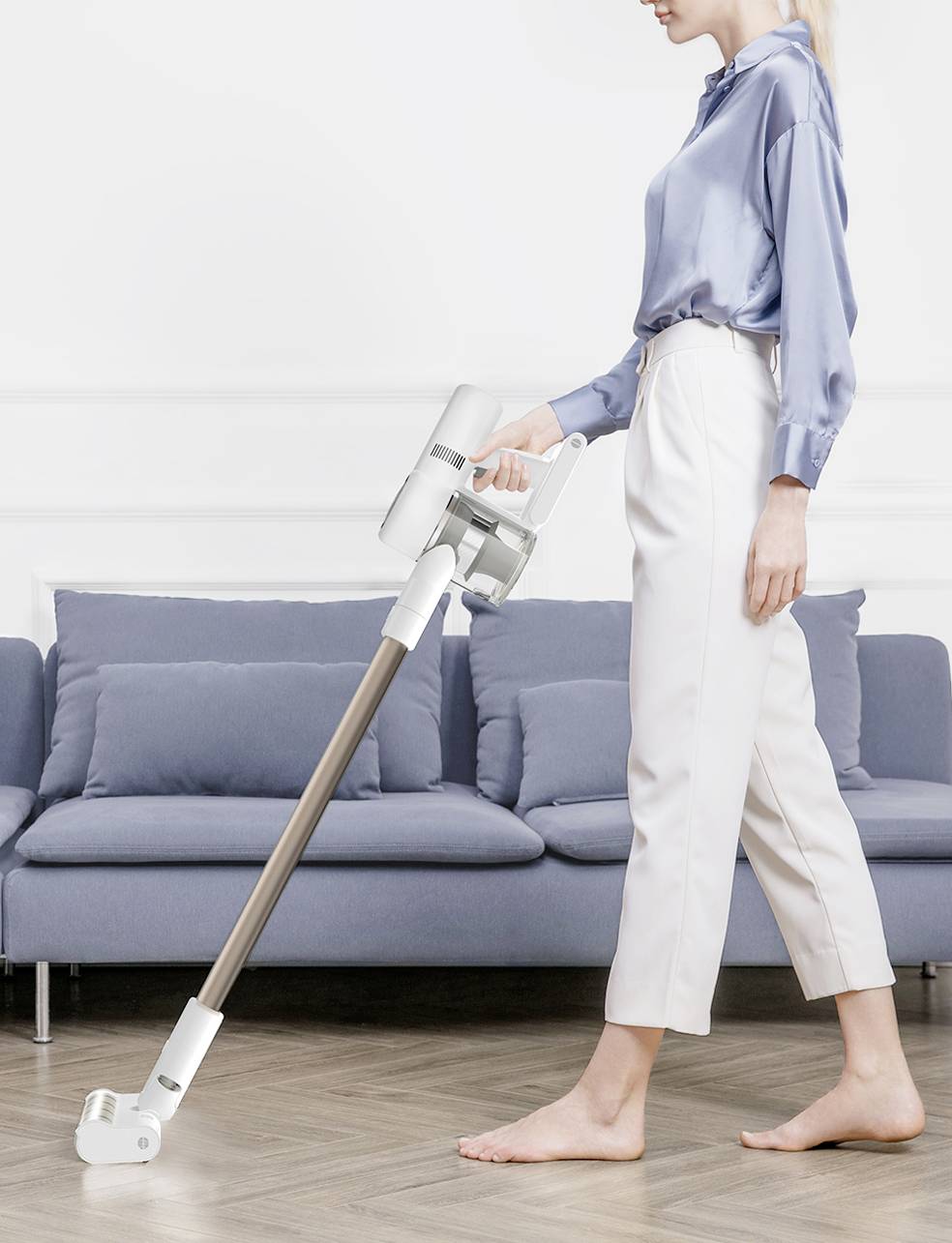 Una persona vestita elegantemente sta usando un aspirapolvere senza fili su un pavimento in legno davanti a un divano blu.