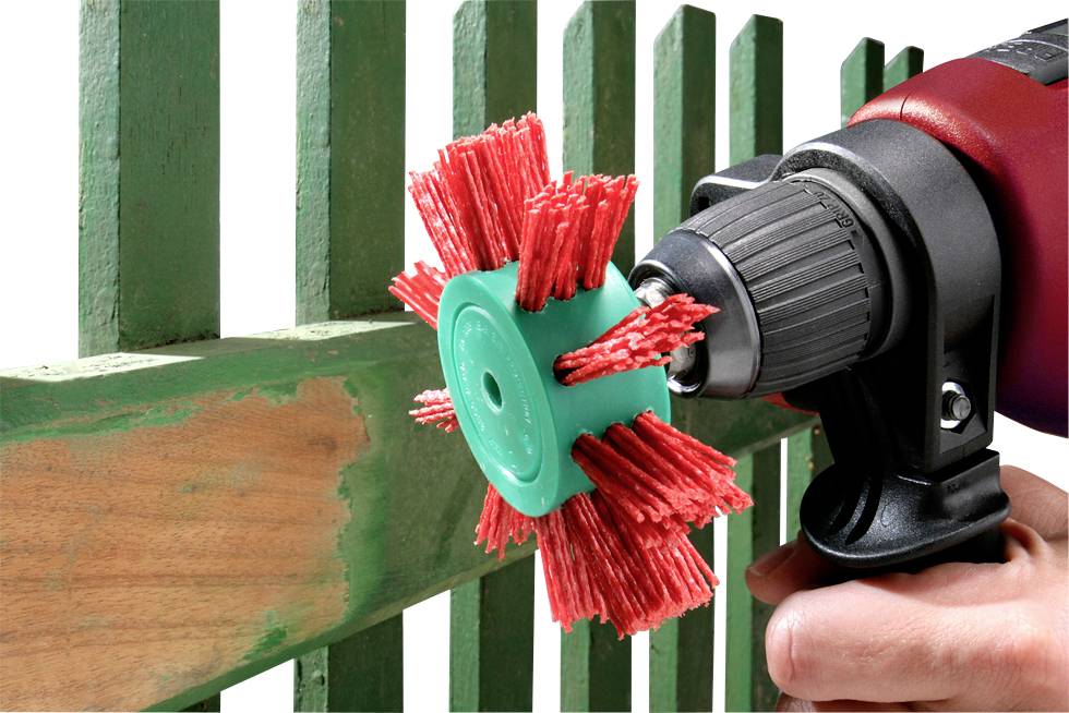 Un trapano con un accessorio abrasivo rosso viene utilizzato per rimuovere la vernice verde da un recinto di legno. La mano tiene saldamente il trapano.
