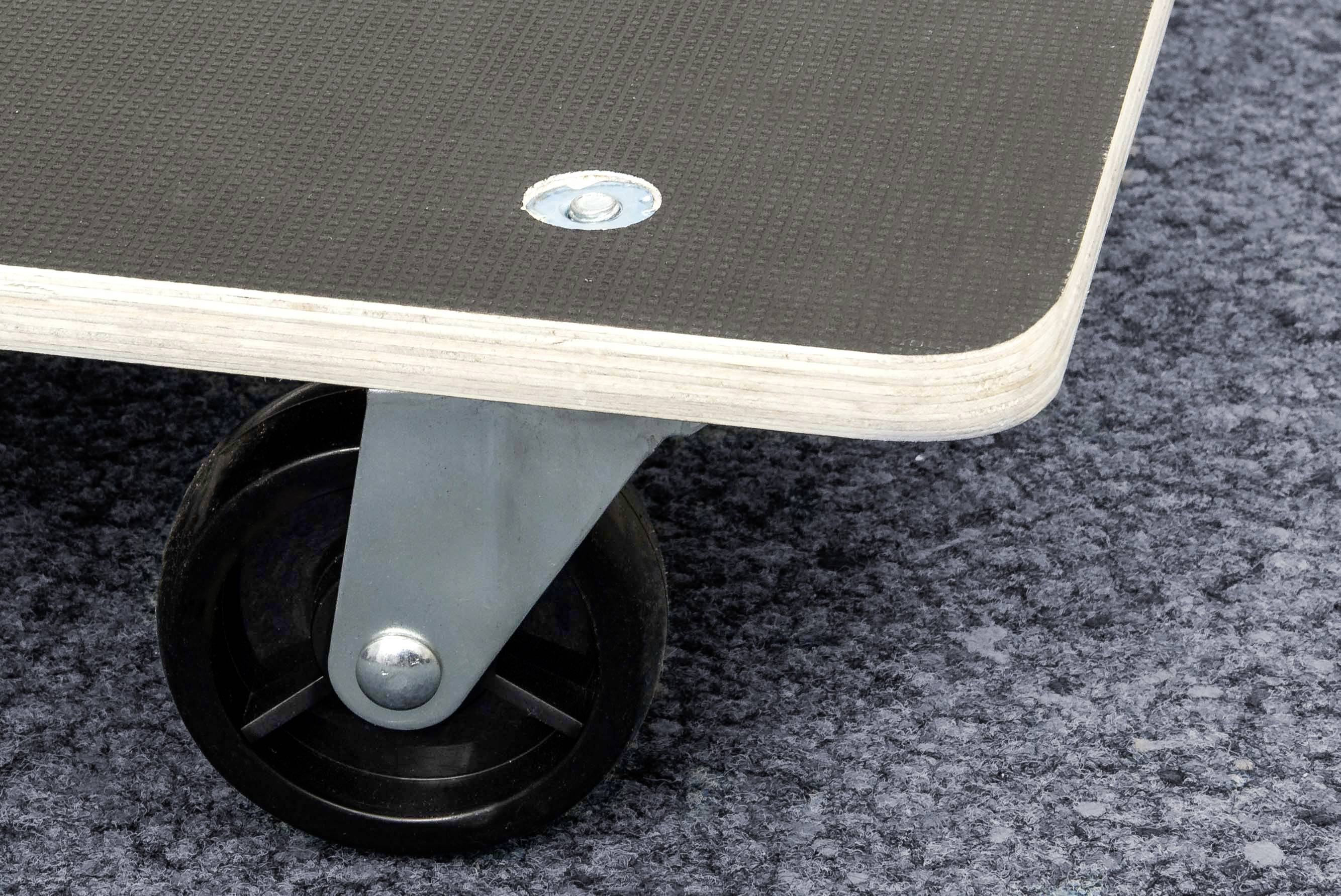 Una ruota nera con supporto in metallo sostiene una tavola rettangolare di legno. Il pavimento è ricoperto da un tappeto grigio.