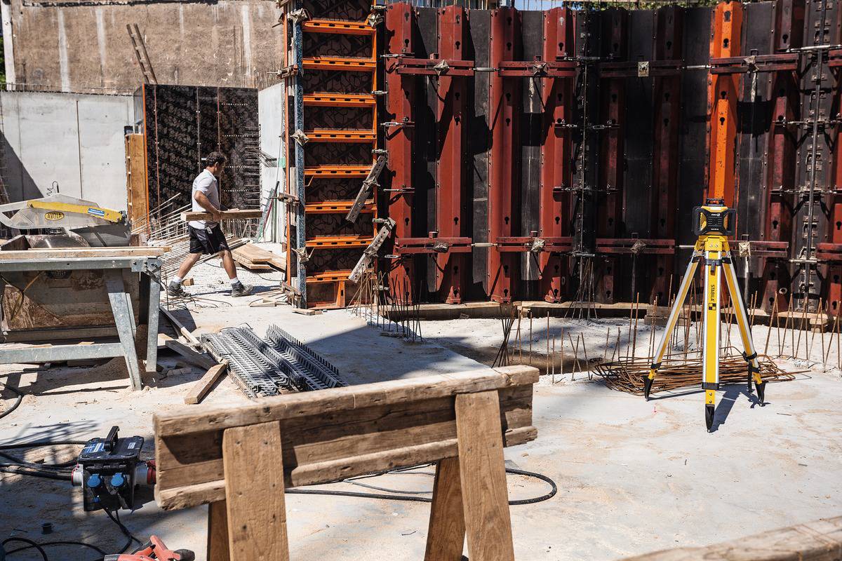 Cantiere con un uomo che monta un telaio in legno. Attrezzi e materiali edili in primo piano. Travi di legno e strumento di misurazione sono visibili.