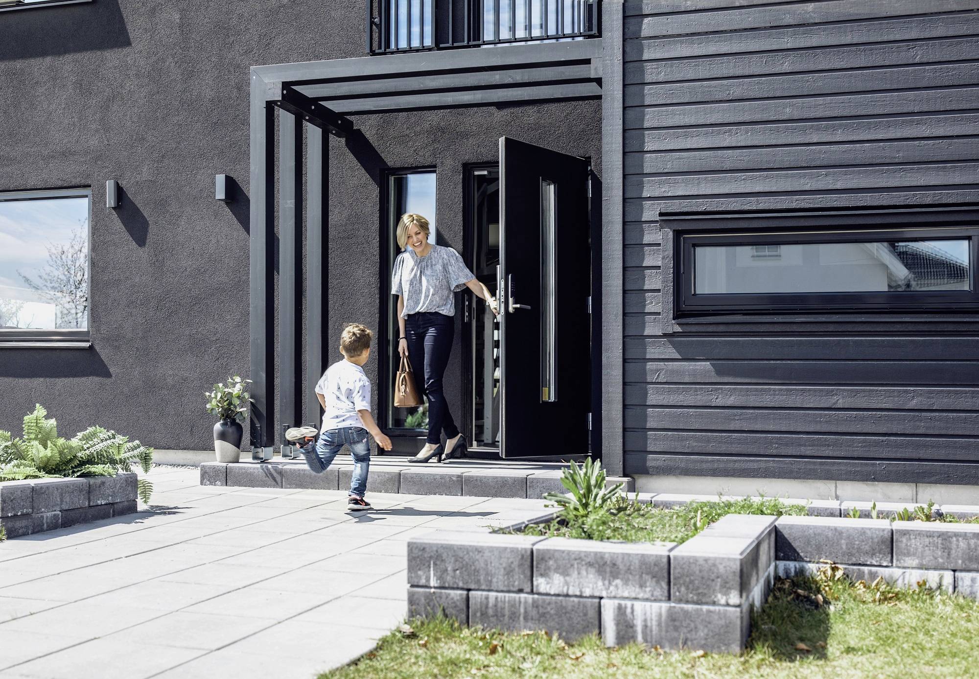 Una donna apre la porta di una casa moderna, mentre un bambino le corre incontro felice. Sullo sfondo sono visibili delle piante.